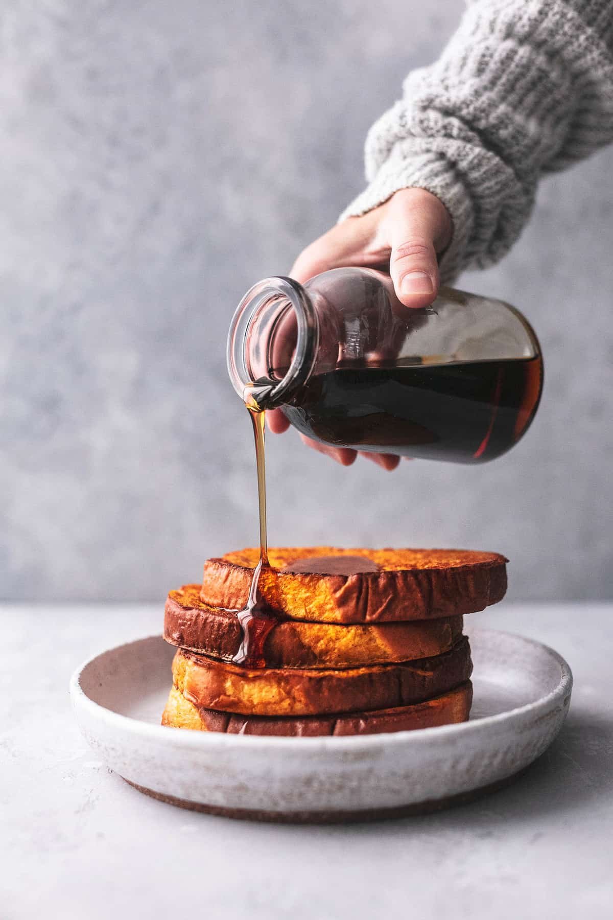 stacked pumpkin French toast on a plate with a hand pouring syrup on top from a jar.