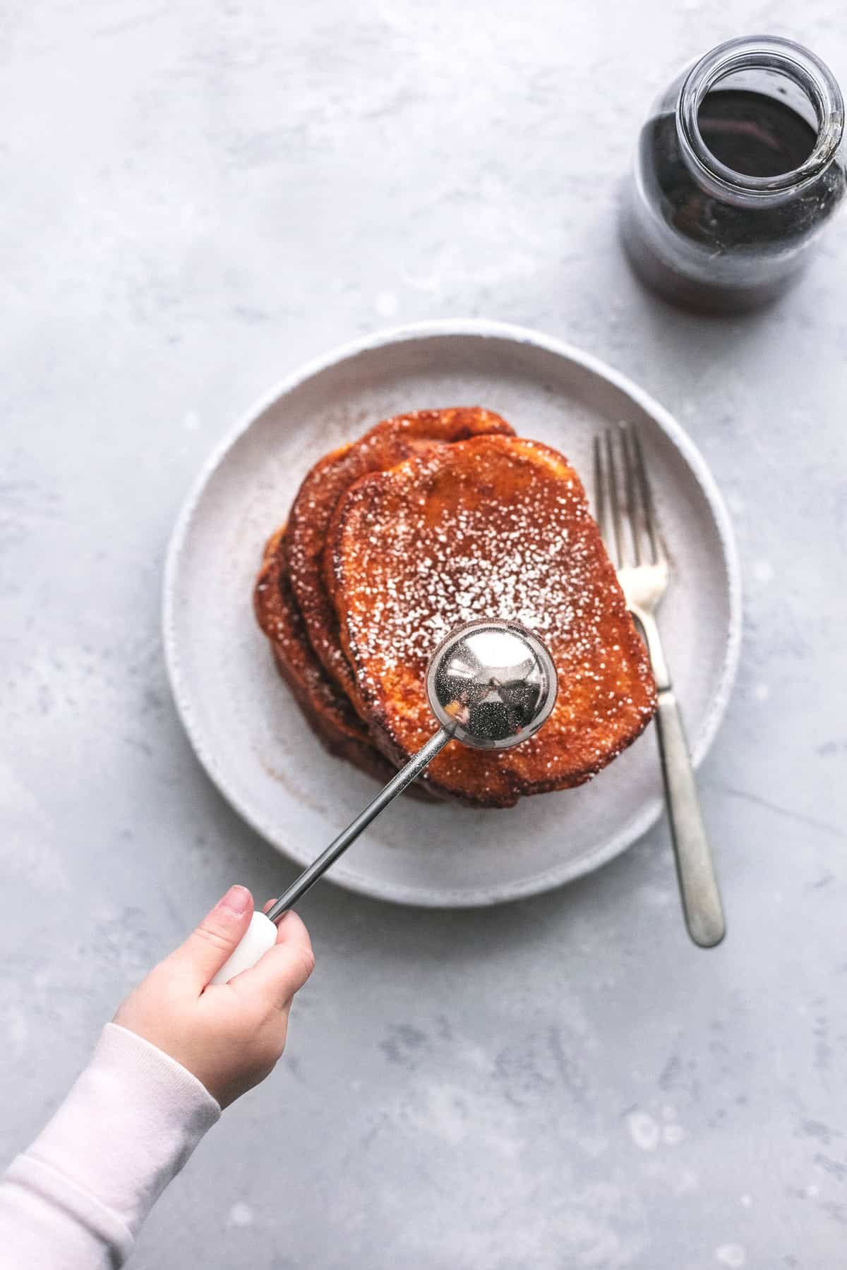 top view of a hand holding a sifter sifting powdered sugar on top of stacked pumpkin French toast with a fork on a plate with a jar of syrup on the side.