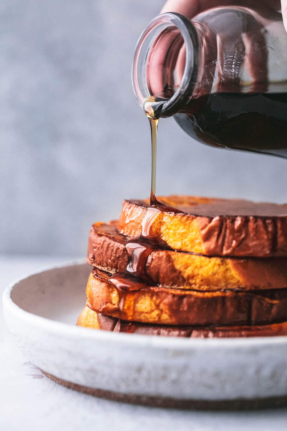 stacked pumpkin French toast on a plate with a hand pouring syrup from a jar on top.