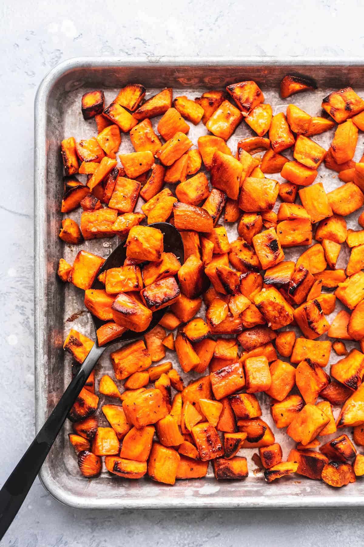 close up top view of roasted sweet potatoes with a serving spoon on a baking sheet.