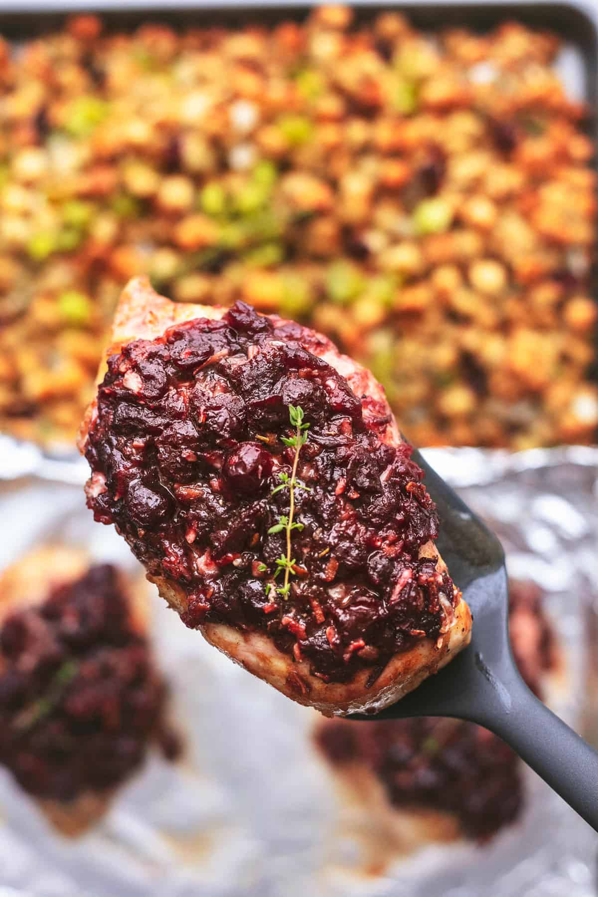 a piece of cranberry chicken on a spatula being held above a sheet pan of more chicken and stuffing.