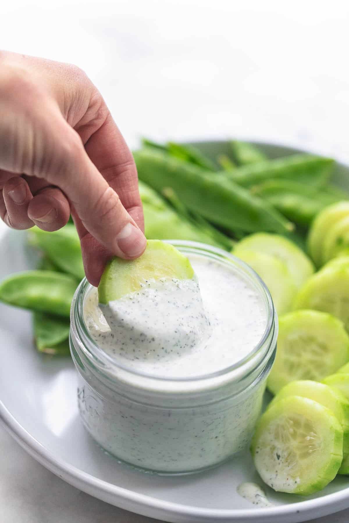 a hand dipping a cucumber slice in a jar of Greek yogurt ranch dressing or dip with more cucumber slices and pea pods on the side all on a plate.