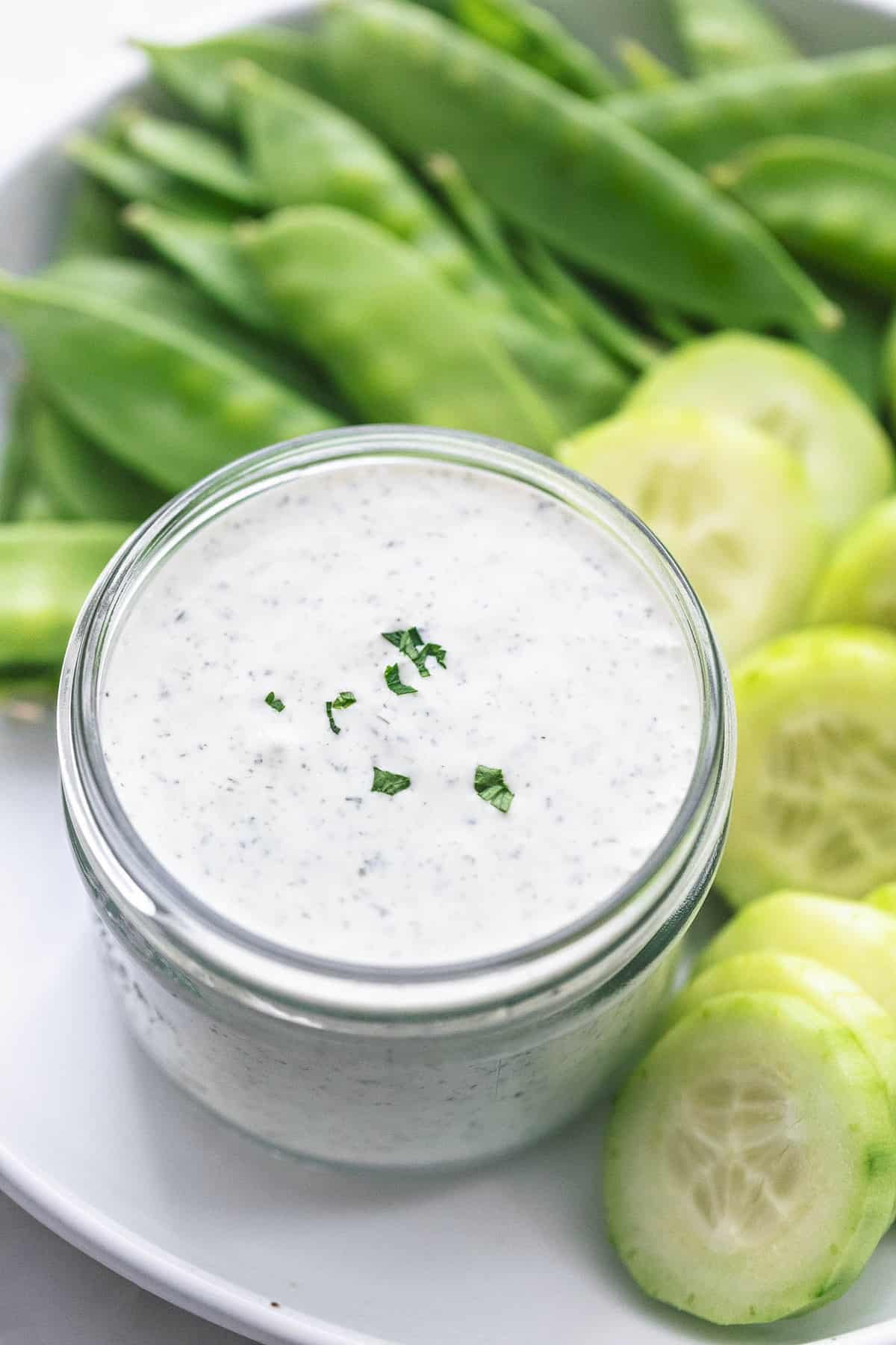 Greek yogurt ranch dressing or dip in a jar with sliced cucumbers and pea pods on the side all on a plate.