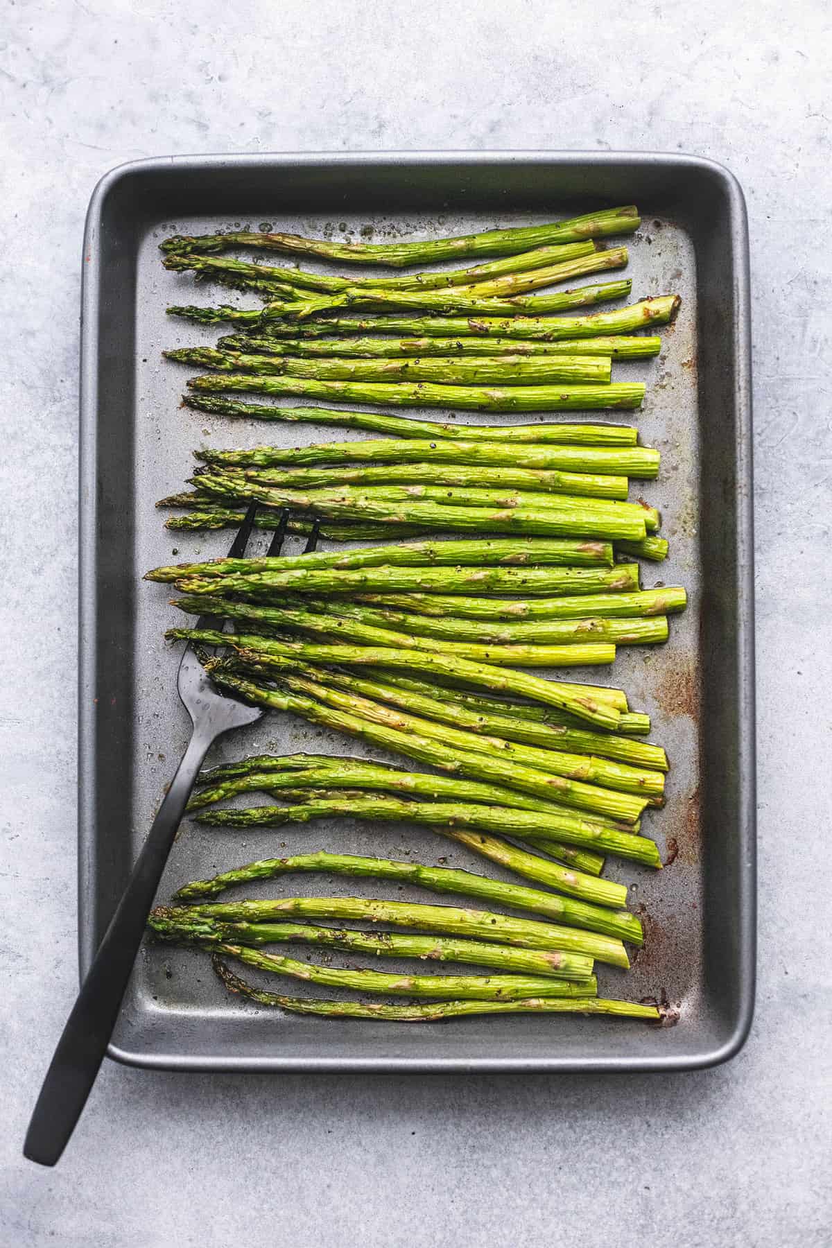 top view of roasted asparagus with a serving fork underneath on the side on a baking sheet.