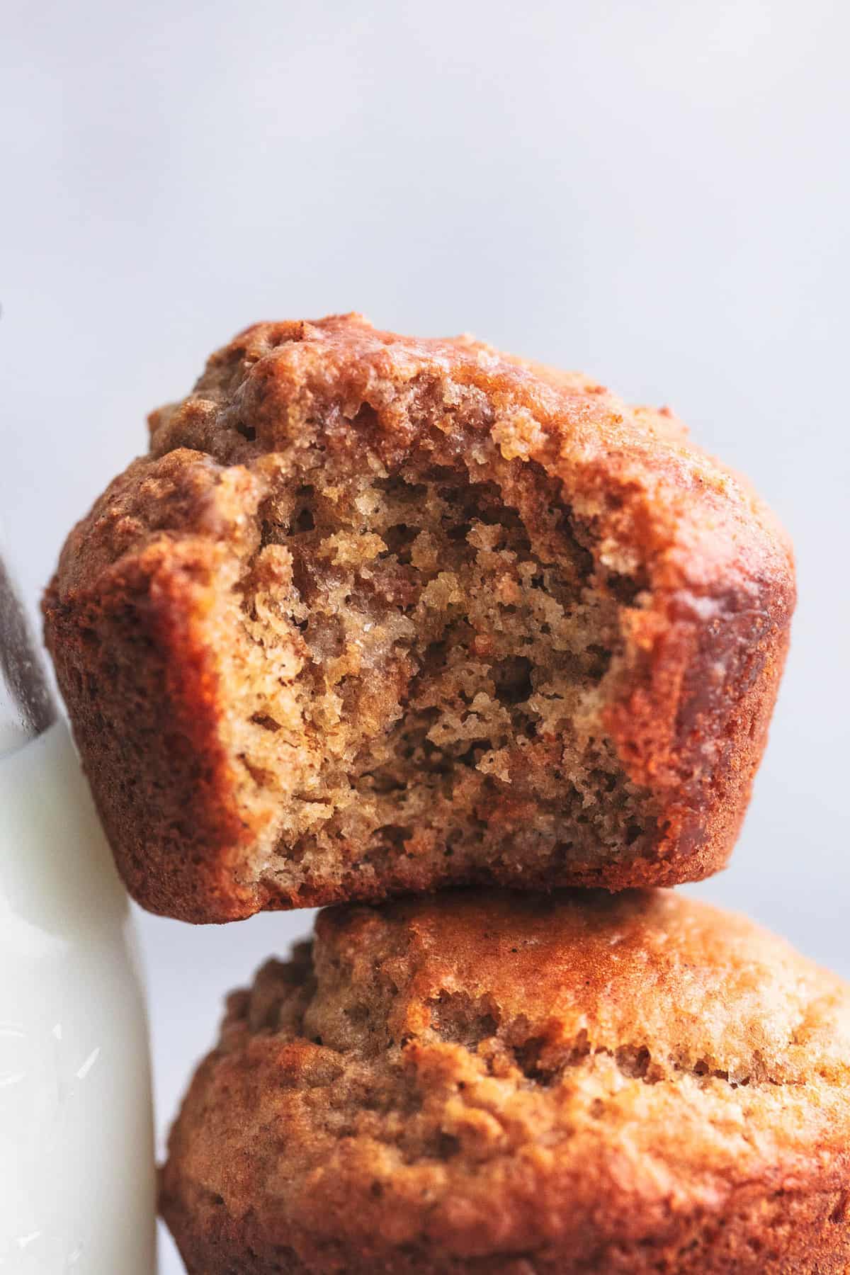 close up of a bran muffin with a bite missing sitting on top of another muffin and leaning on a glass pitcher of milk.