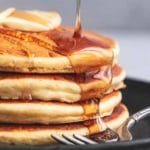 up close syrup pouring onto stack of pancakes on a plate with a fork