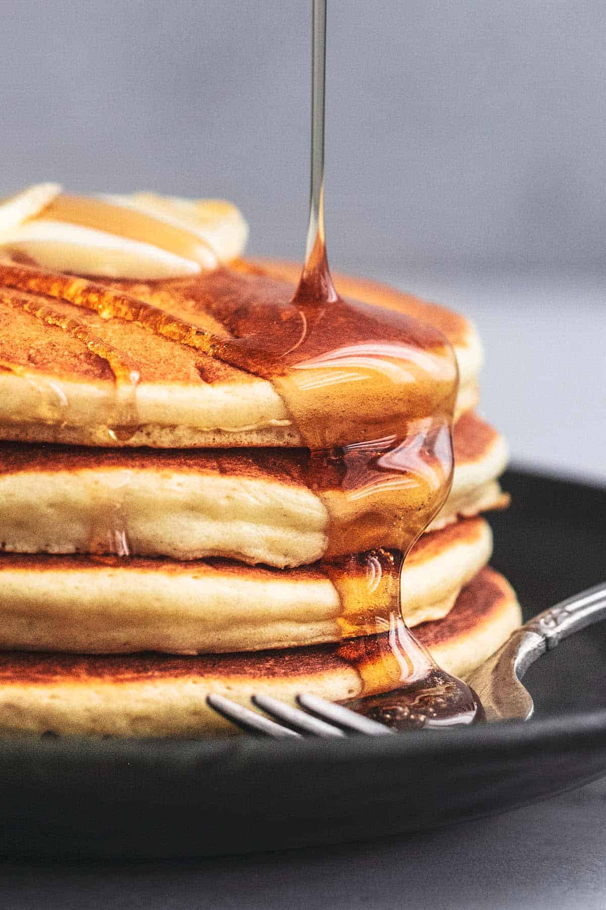 close up of syrup pouring onto stack of fluffy buttermilk pancakes on a plate with a fork.