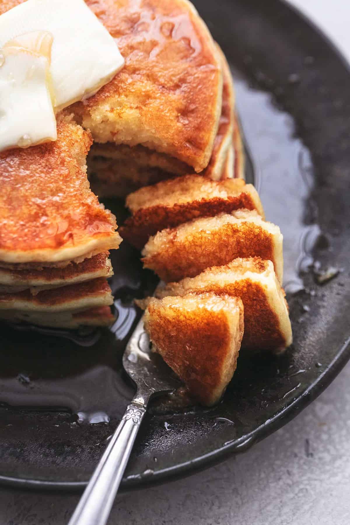 close up top view of a bite of fluffy buttermilk pancakes with the rest of the pancakes on the side with syrup and butter on black plate.