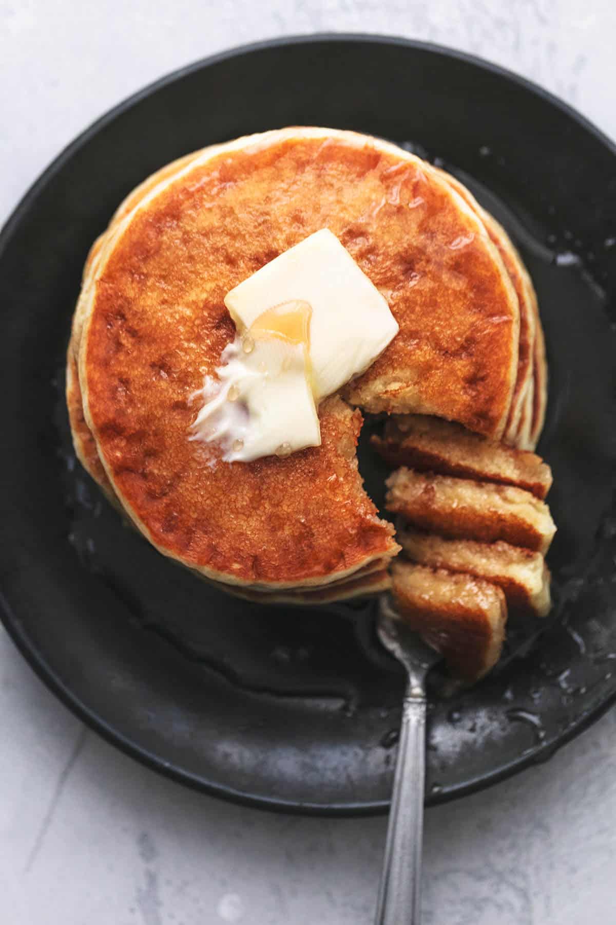 top view of a stack of fluffy buttermilk pancakes topped with butter and syrup with a bite on the side on a fork all on a plate.