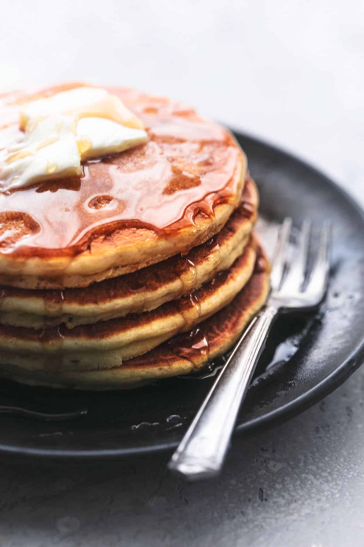 close up of fluffy buttermilk pancakes topped with butter and syrup on a plate with a fork.