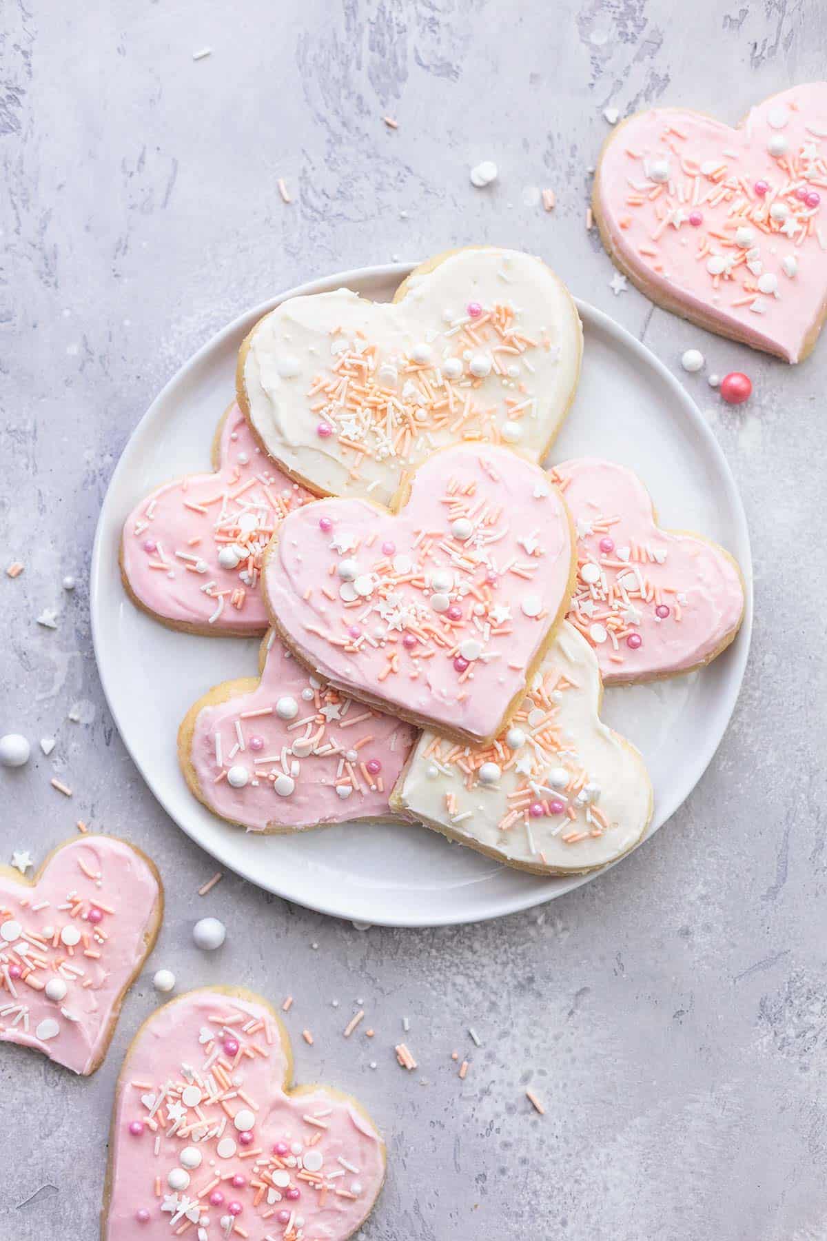 top view of heart shaped sugar cookies with sprinkles on a white plate with more cookies on the side.