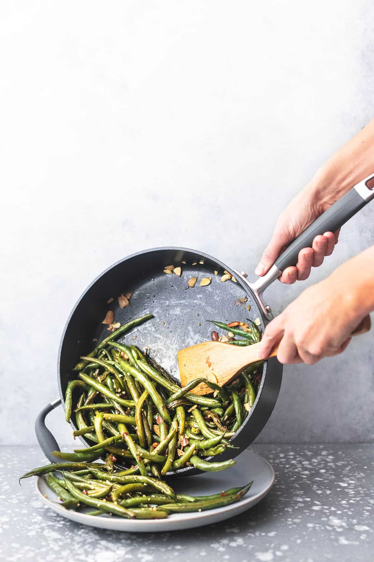 sautéed green beans with garlic and almonds being scooped out of a skillet onto a plate with a wooden spatula.