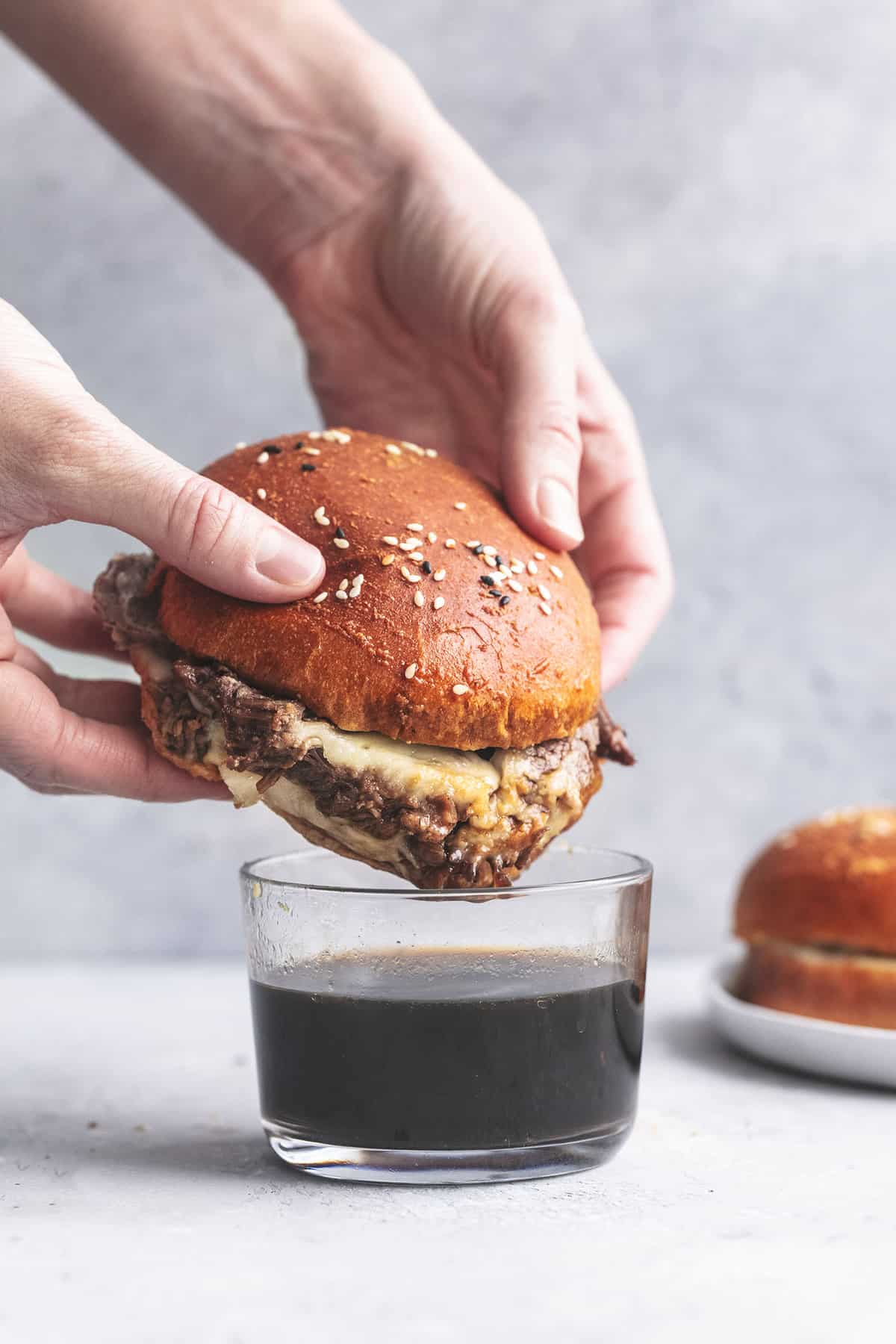 a hand dipping a french dip sandwich in au jus sauce in glass.