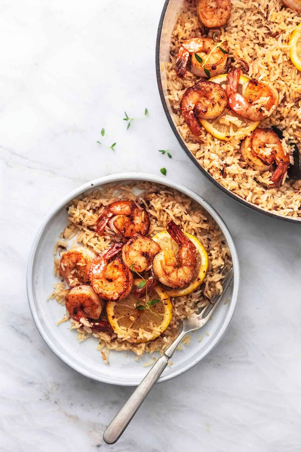 top view of lemon herb shrimp and rice skillet with lemon slices on a plate with a fork with more shrimp and rice in a skillet on the side.