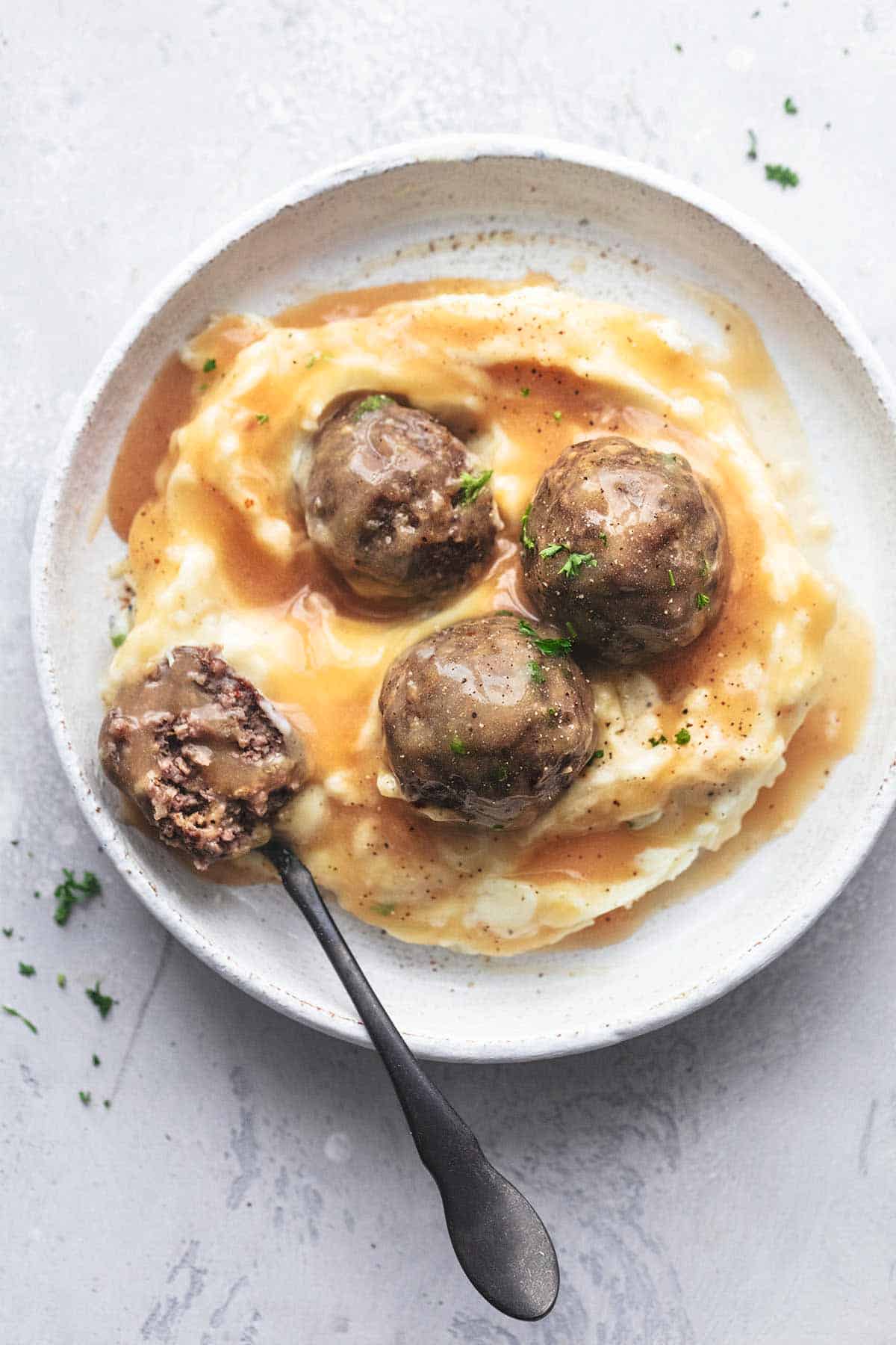 top view of baked meatballs on mashed potatoes and gravy with half of a meatball on a fork on the side all on a plate.