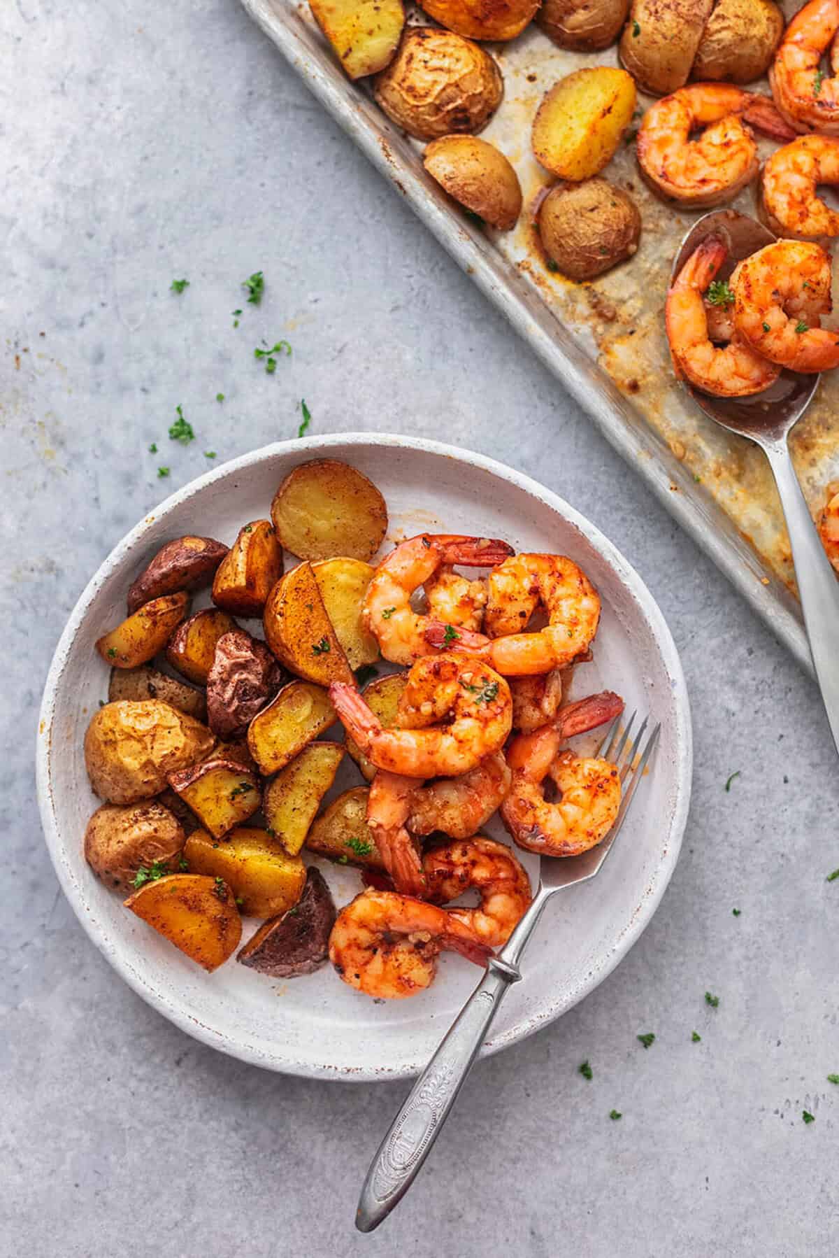 plate of potatoes with shrimp and fork next to a sheet pan with serving spoon.