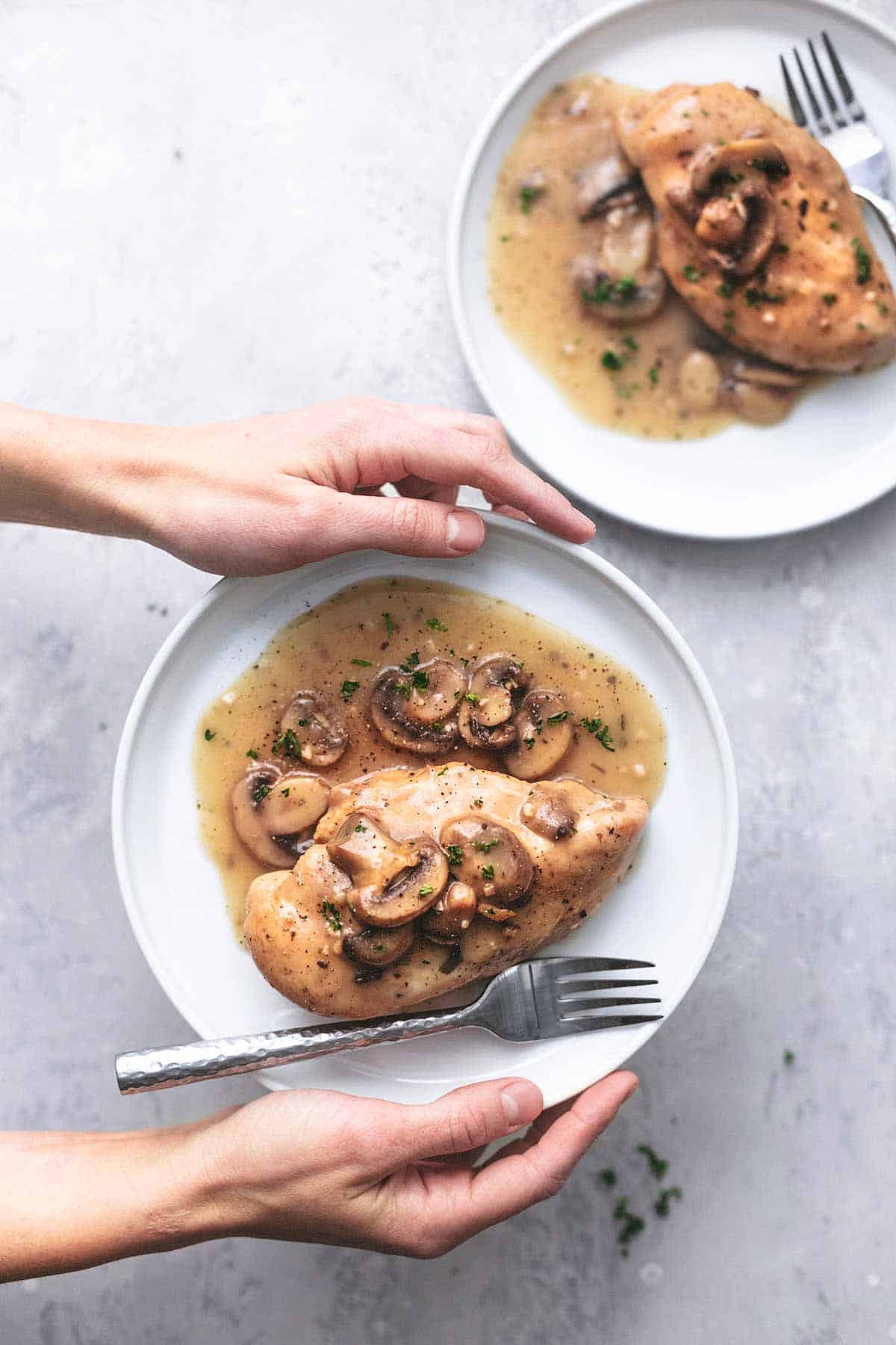 hands holding chicken with gravy and mushrooms on a white plate with a fork