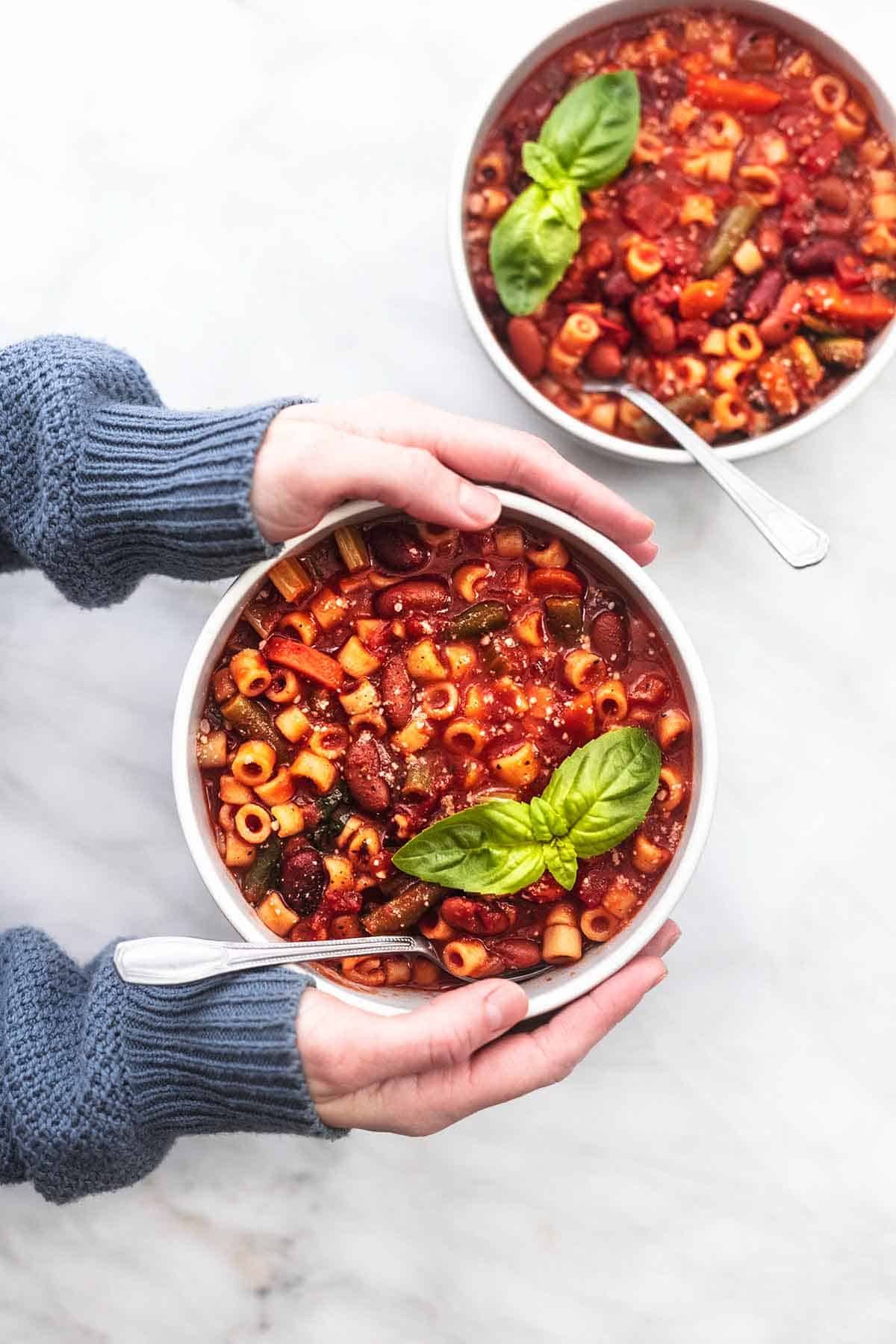 hands holding one bowl of minestrone soup with second bowl of soup on table