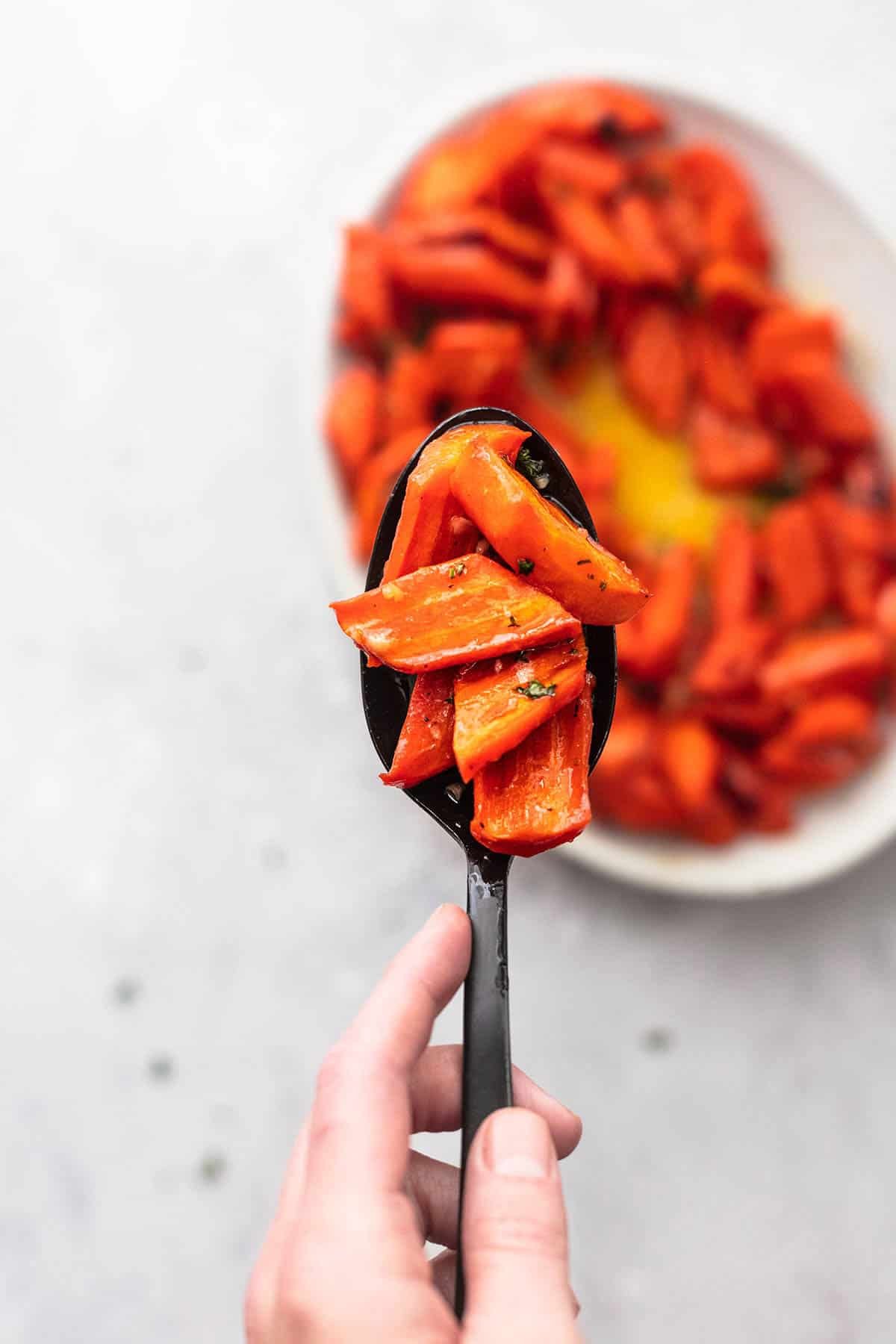 top view of a hand holding roasted honey garlic glazed carrots on a spoon above a plate of carrots.