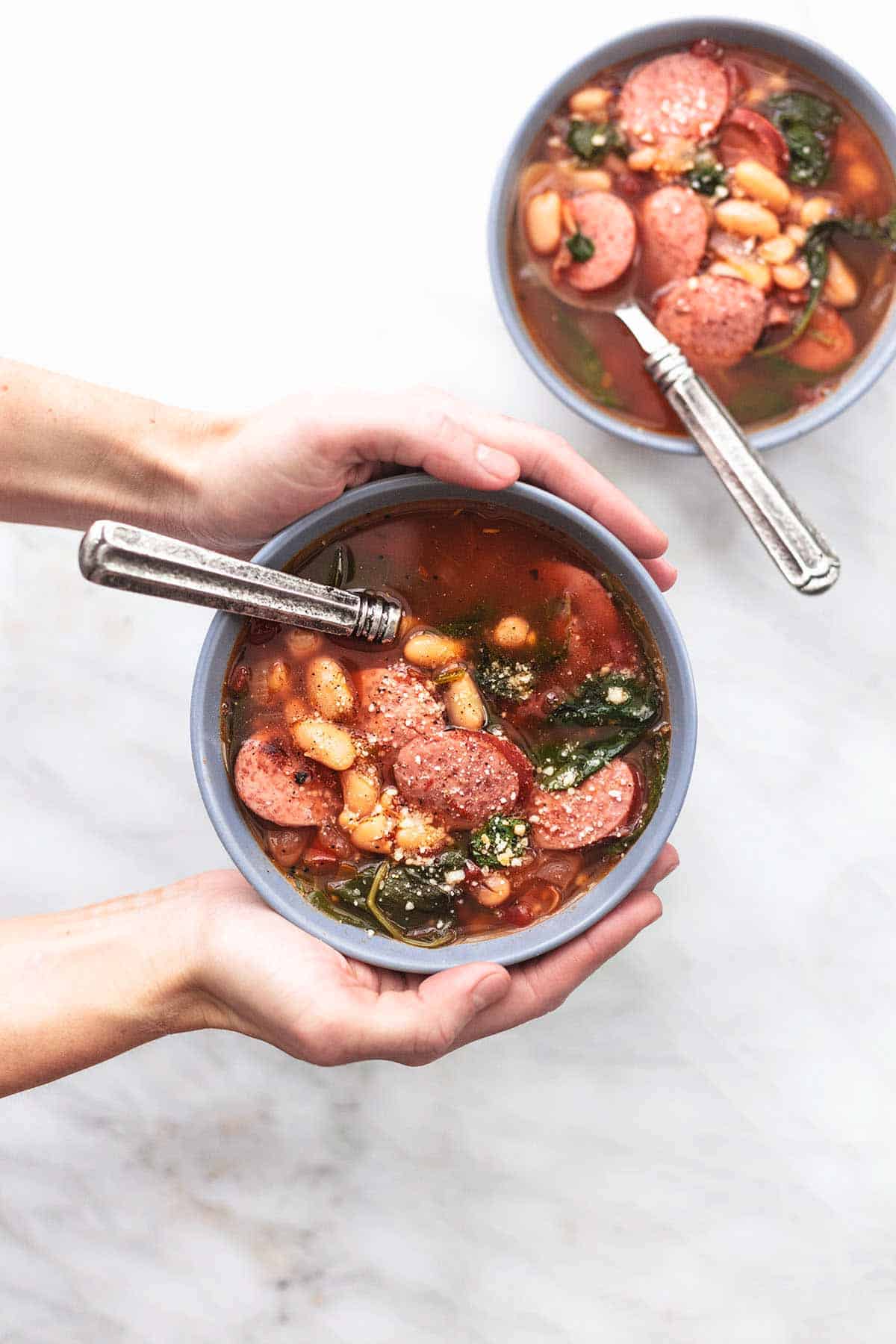 top view of hands holding a bowl of sausage and white bean soup with a spoon with another bowl of soup with a spoon on the side.