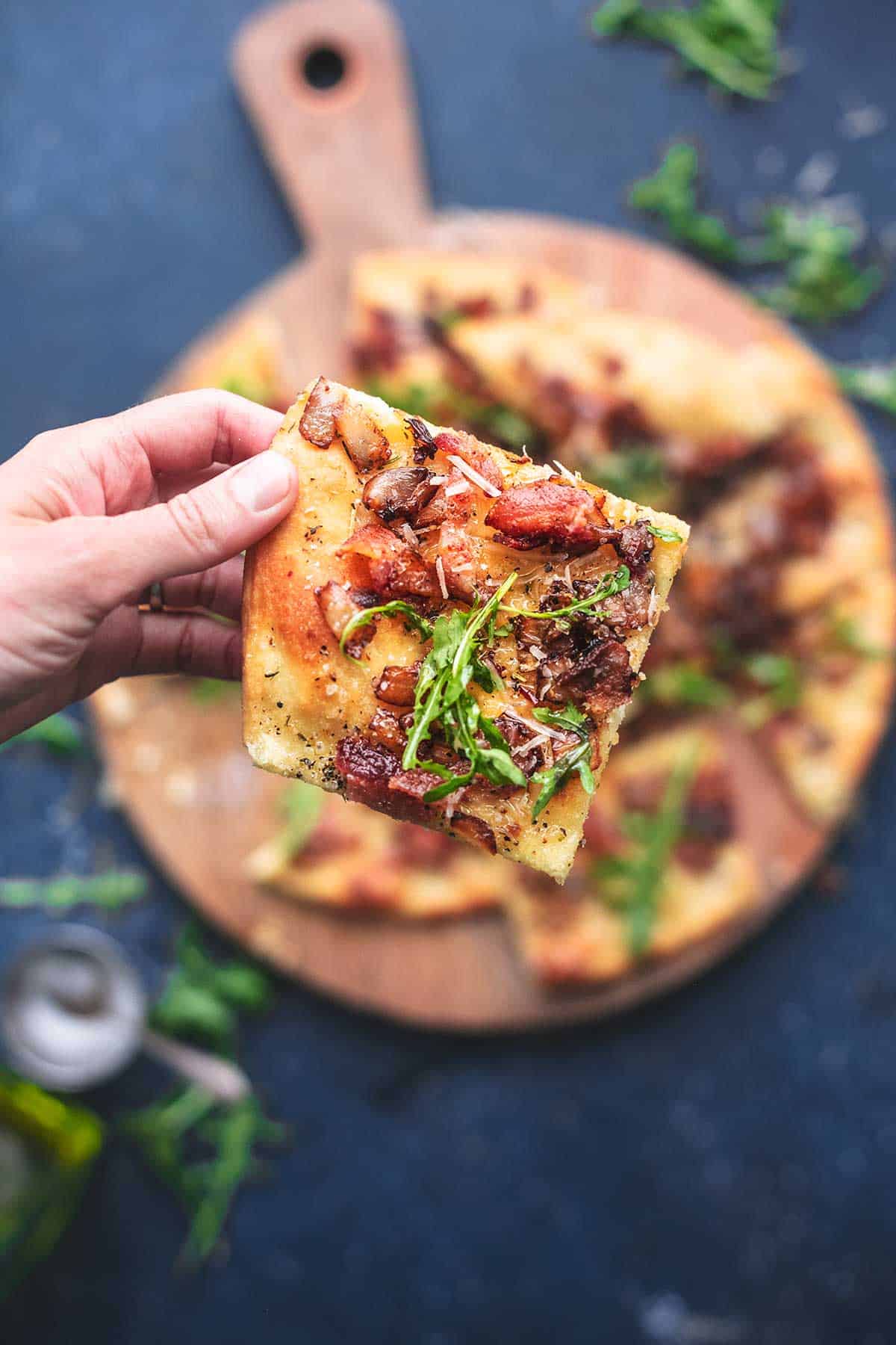 top view of a hand holding a piece of caramelized onion and bacon flatbread.