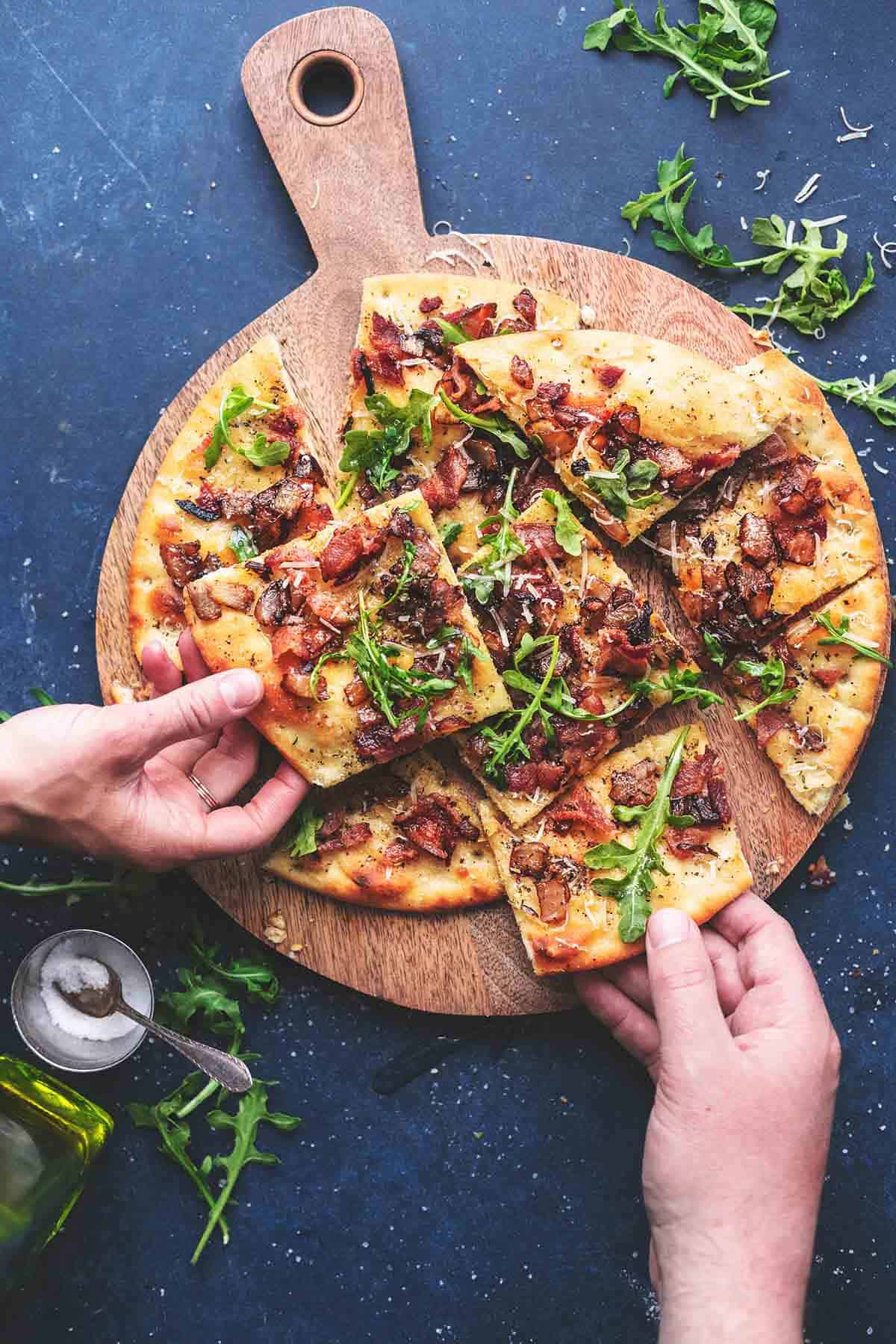 top view of hands grabbing pieces of caramelized onion and bacon flatbread from a wooden serving board.