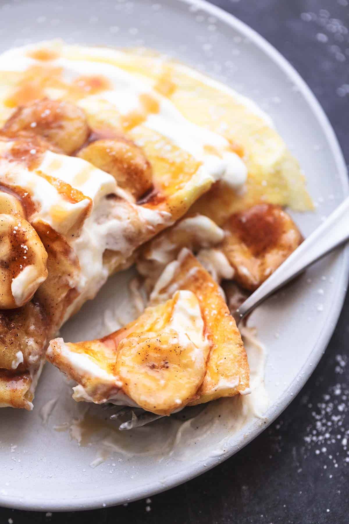 close up of a bite of crepes with banana crepe filling on a fork with the rest of the crepes on the side all on a plate.