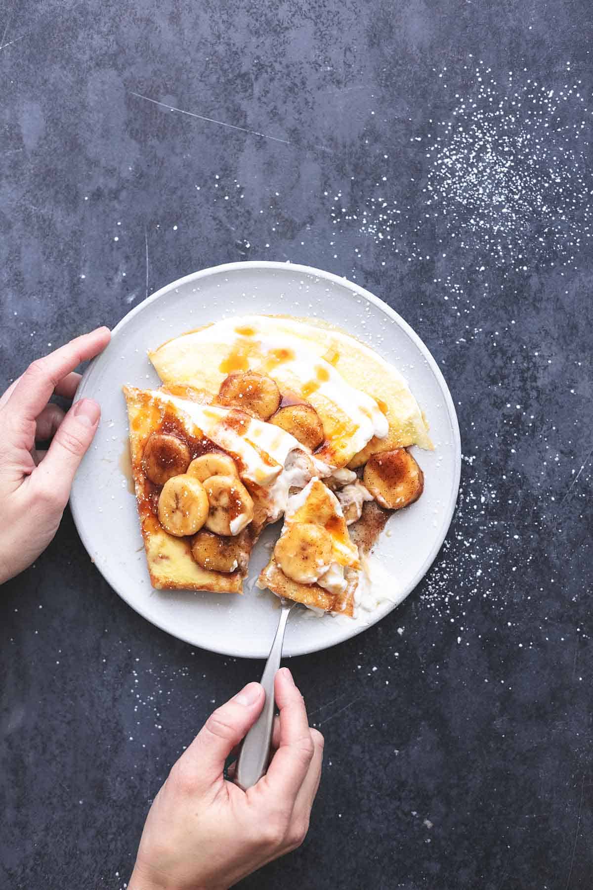 top view of a hand holding the side of a plate of crepes with banana crepe filling with another hand holding a fork under a bite.