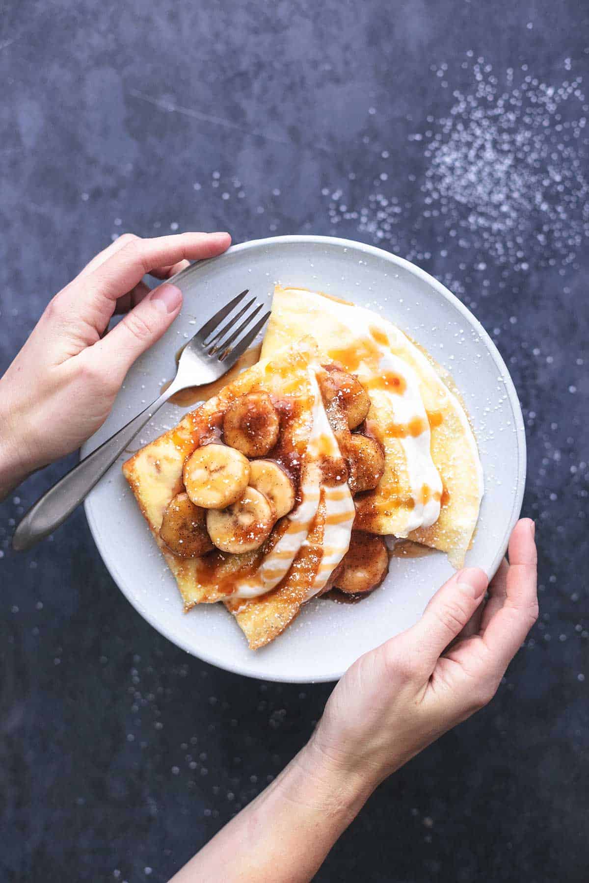 top view of hands holding a plate with crepes with banana crepe filling and a fork on it.