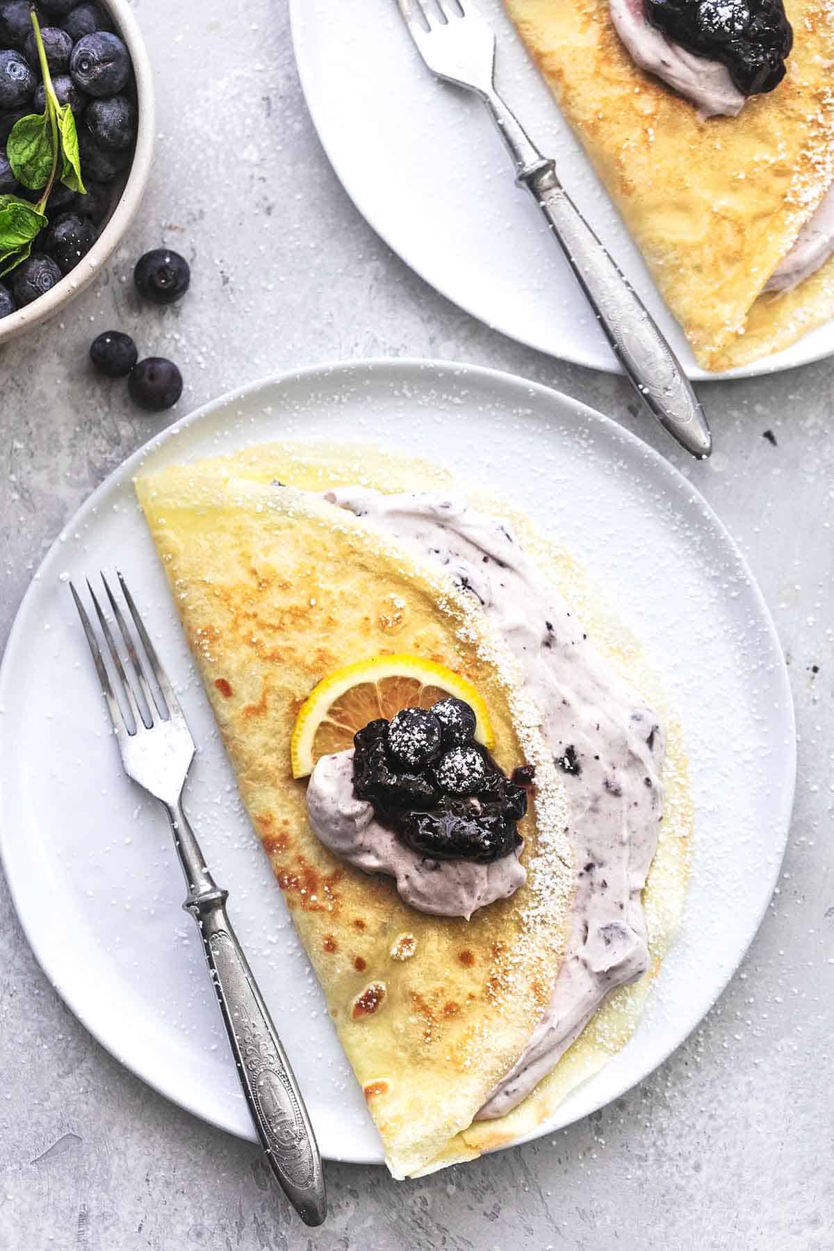 top view of a crepe with blueberry filling with a fork on a plate with a bowl of blueberries and another plate on the side.