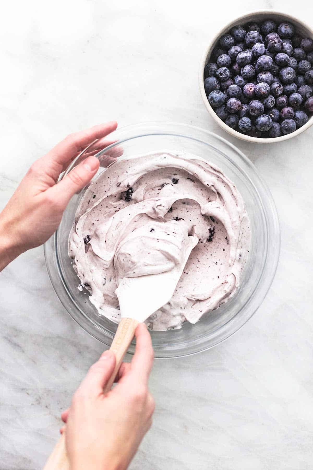 top view of a hand holding the side of a glass bowl of blueberry crepe filling and the other hand holding a spatula in the bowl with a bowl of blueberries on the side.