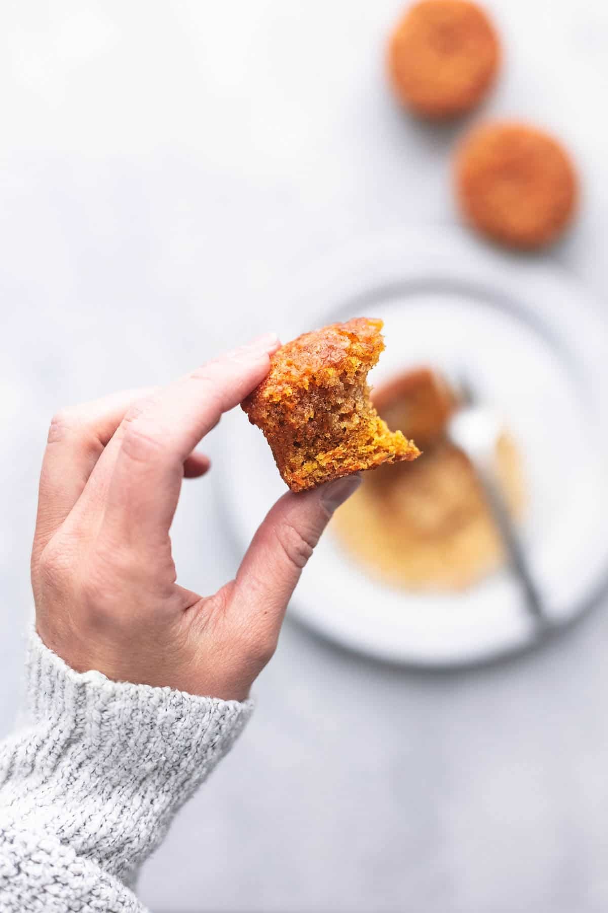 top view of a hand holding bite of a carrot cake muffin above a plate with half of a muffin and a fork on it with more muffins on the side.