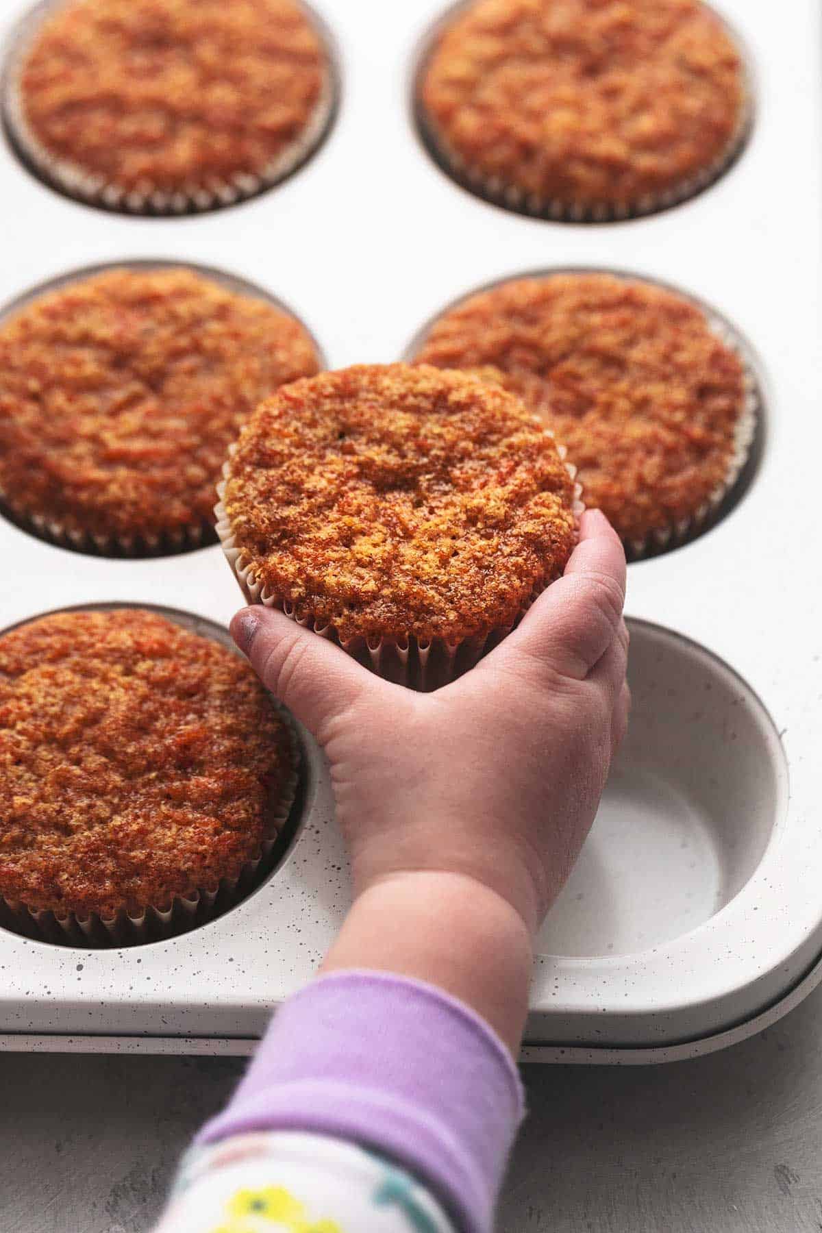 a child's hand holding a carrot cake muffin on a muffin tin with more muffins.