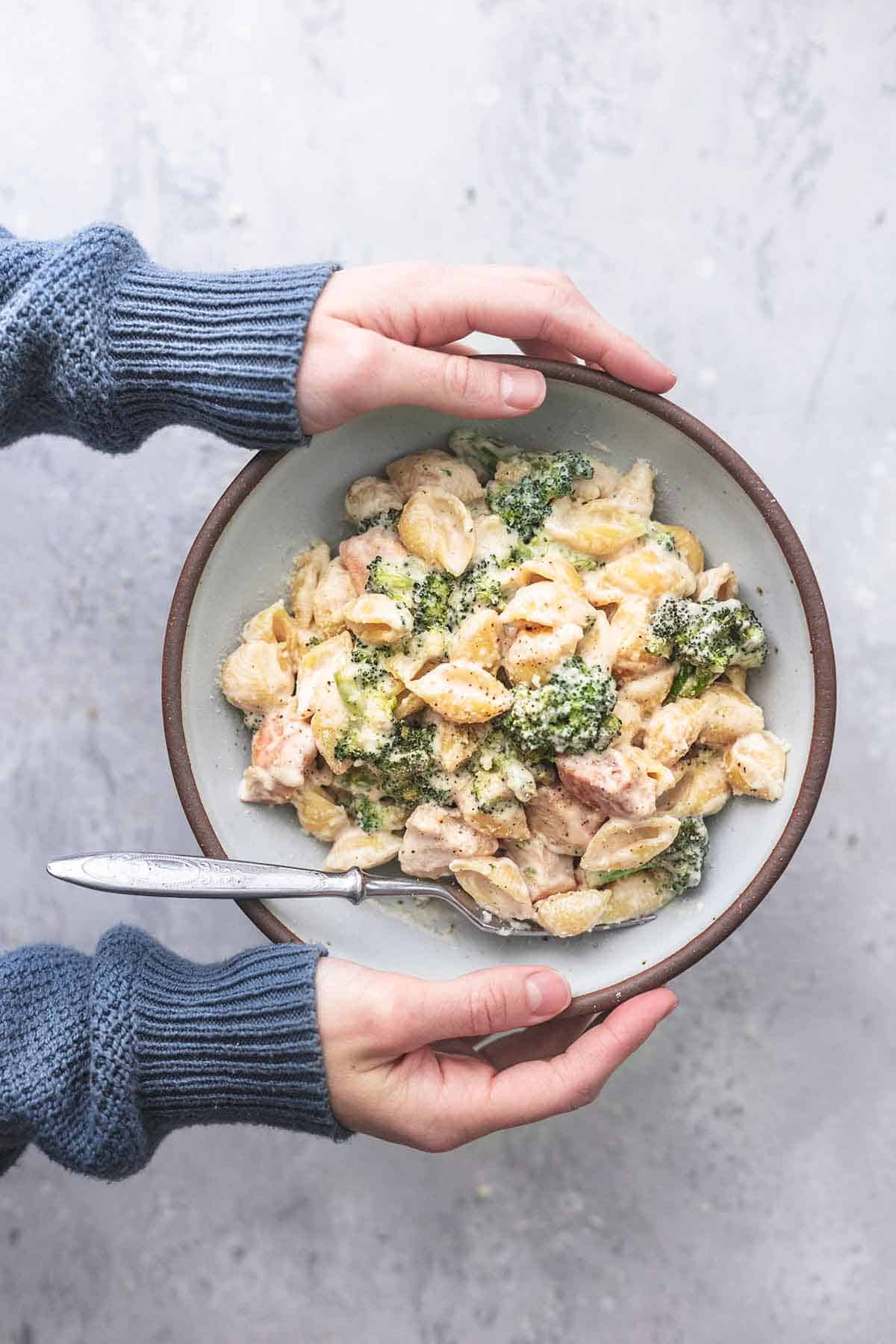 top view of hands holding bowl of chicken alfredo with broccoli.