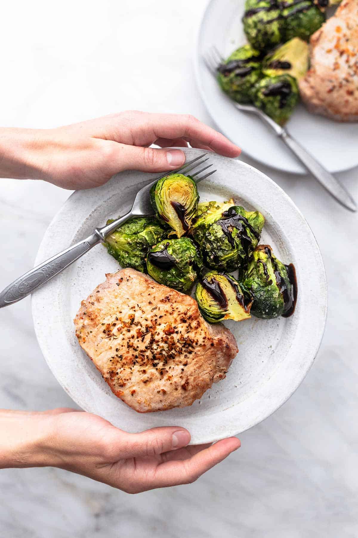 top view of hands holding plate of pork chops and brussels sprouts with a fork with another plate on the side.