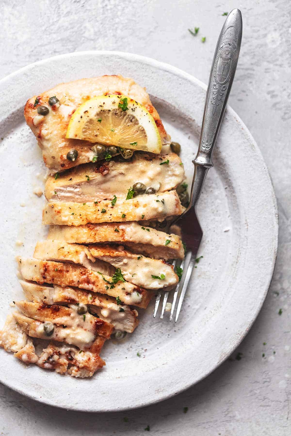 overhead sliced lemon chicken and capers with fork on white plate