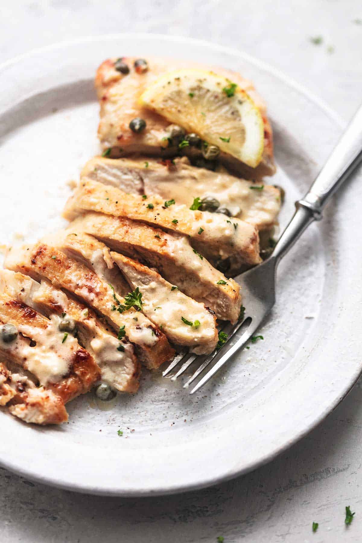 close up of a piece of creamy lemon chicken piccata sliced up on a plate with a fork.