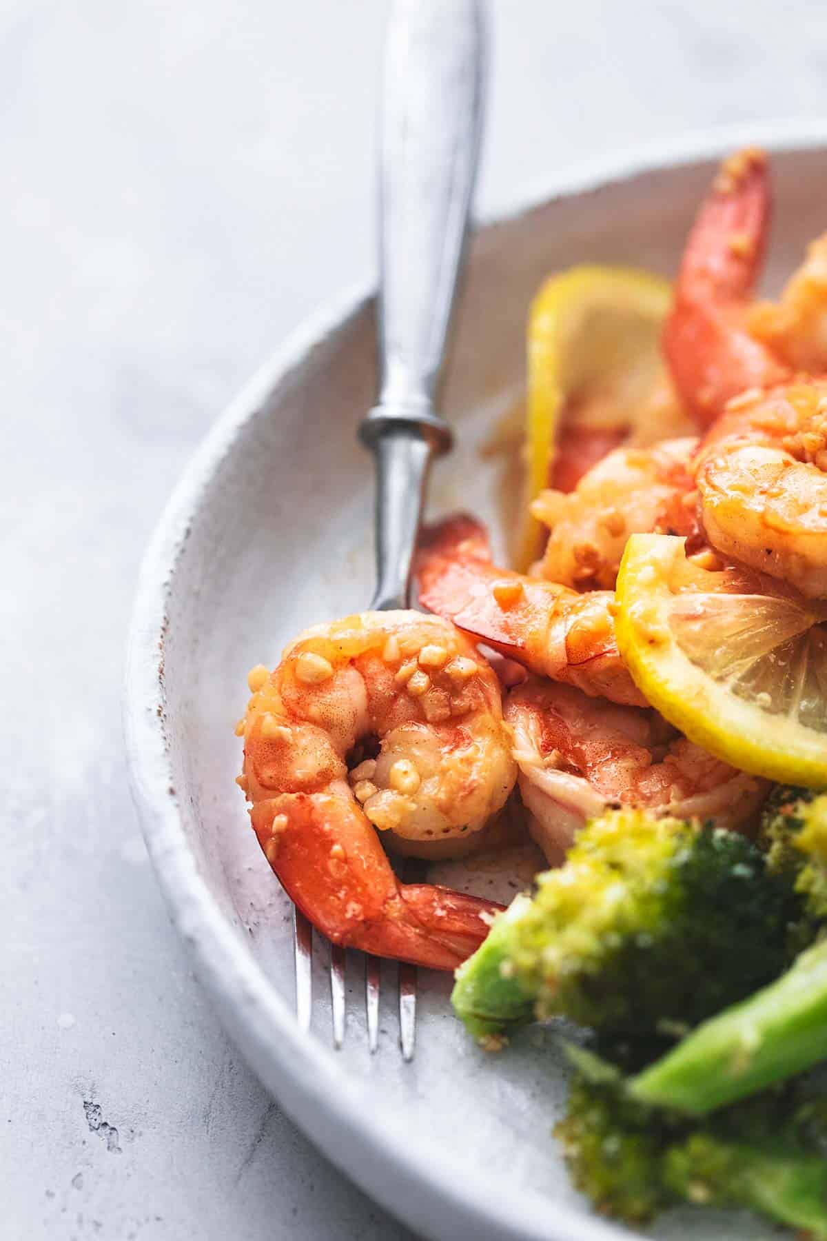 close up of a piece of shrimp on a fork with more sheet pan shrimp scampi and broccoli on a plate.