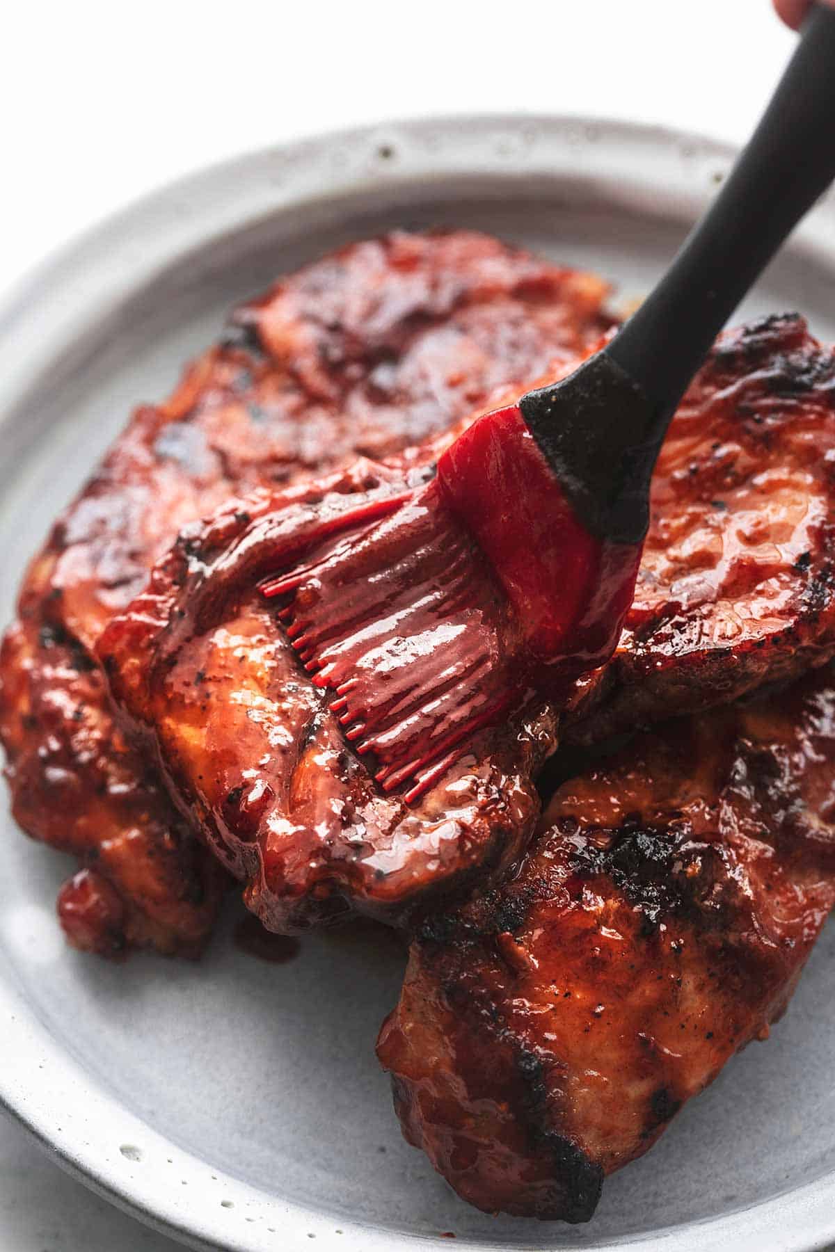 close up of a brush basting bbq sauce onto grilled pork chops on a plate.