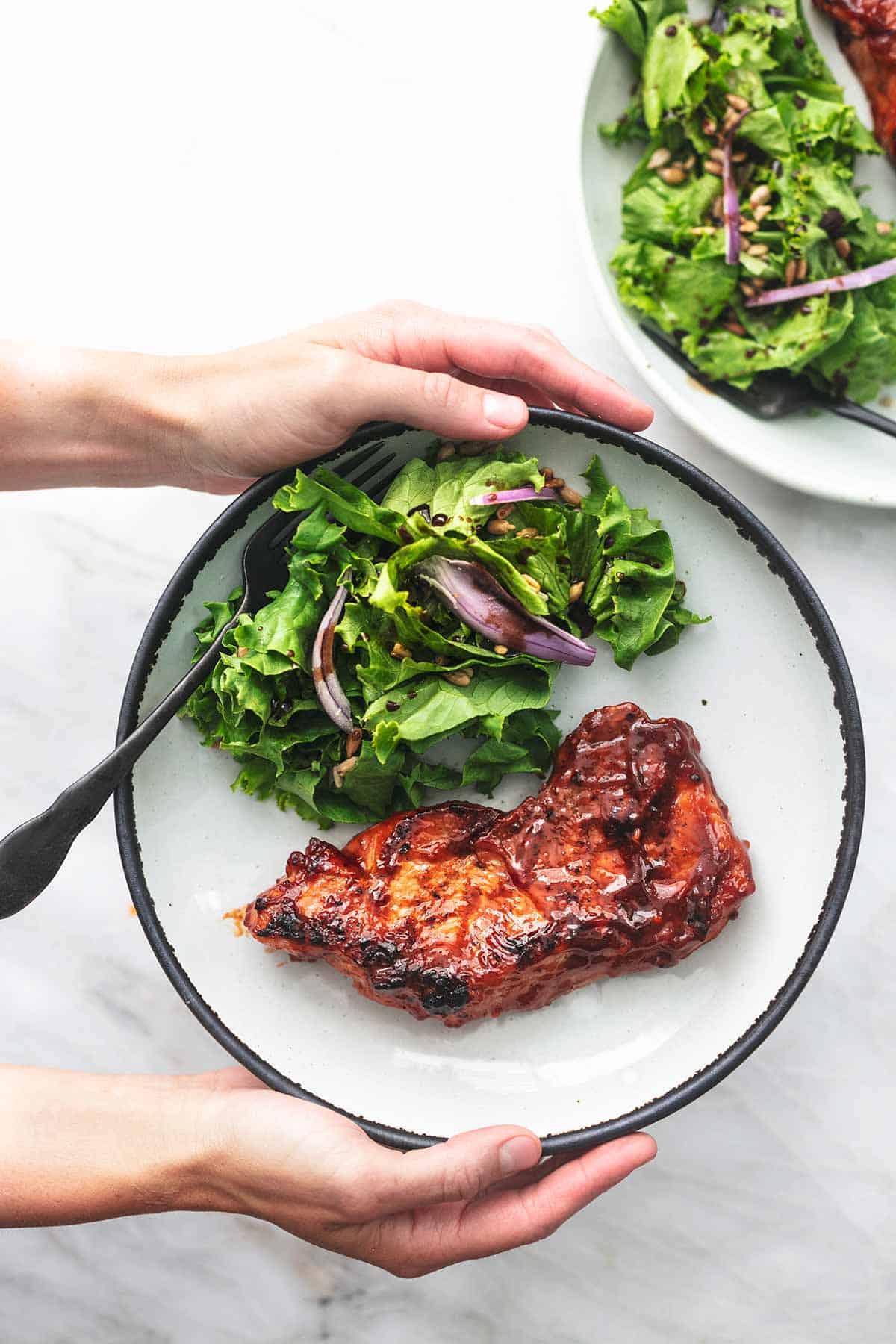 top view of hands holding a plate with a grilled bbq pork chop, salad, and a fork on it.