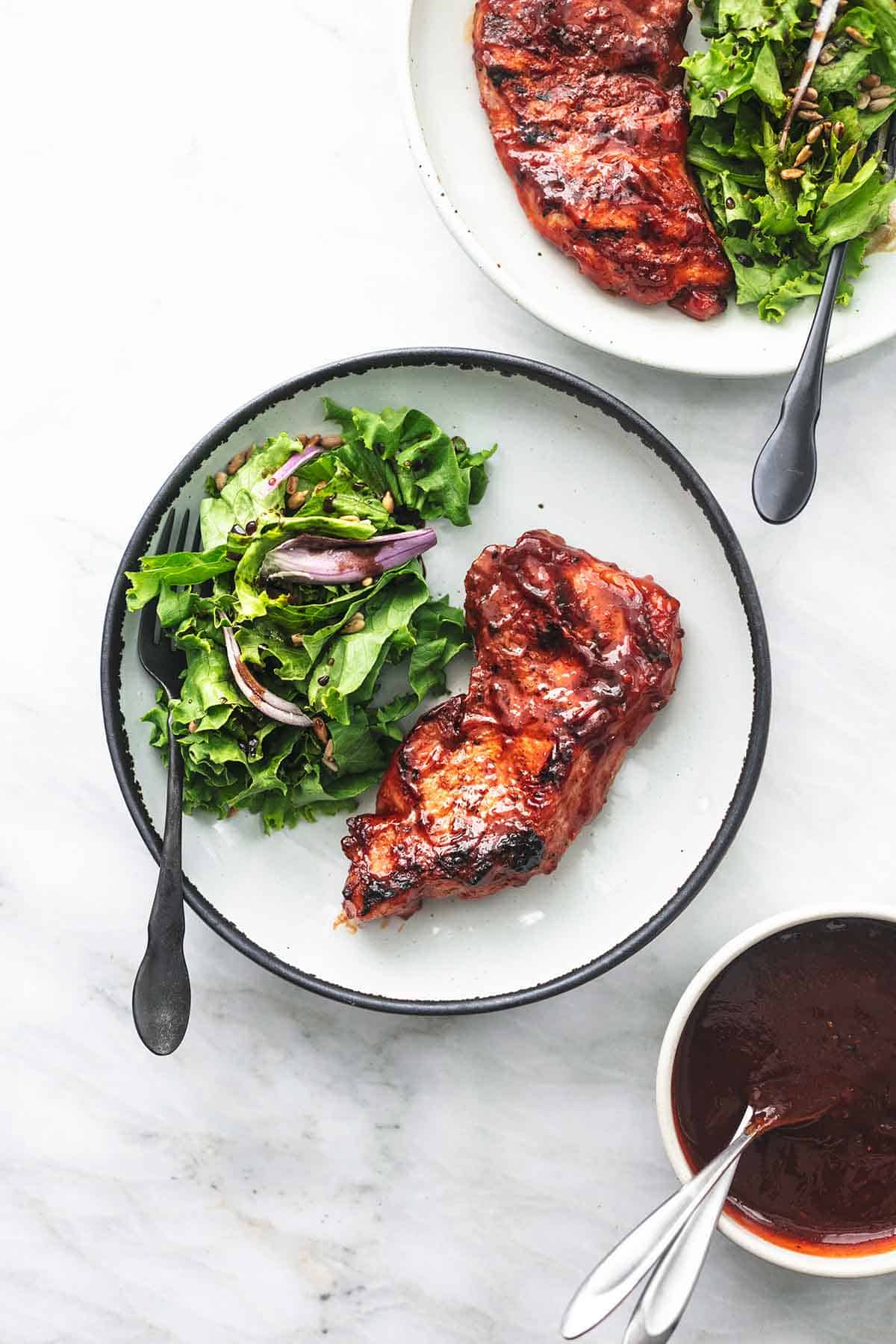 top view of two plates with grilled bbq pork chops, salad and forks on them with a bowl of bbq sauce on the side.