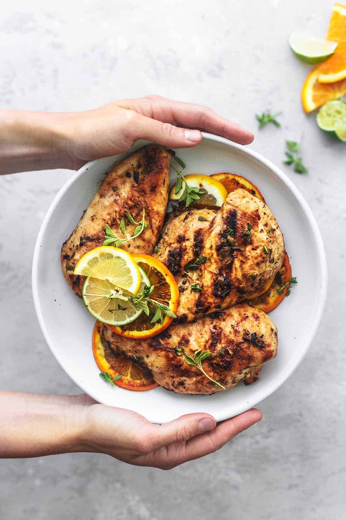 top view of hands holding a white plate of citrus dijon grilled chicken (marinade) with lemon and orange slices with lime and orange slices on the side.