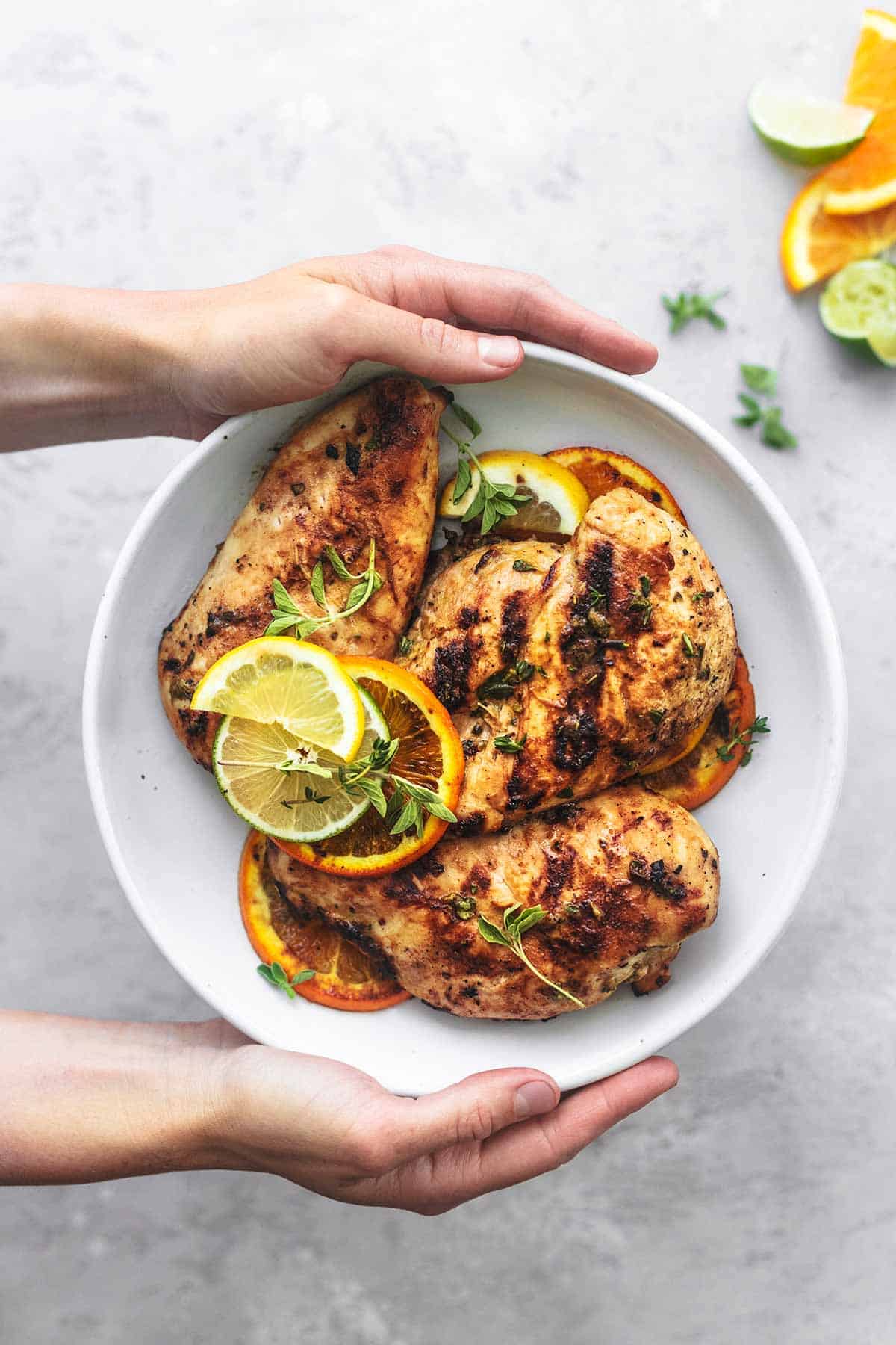 overhead view of hands holding white plate of grilled chicken with citrus fruit slices