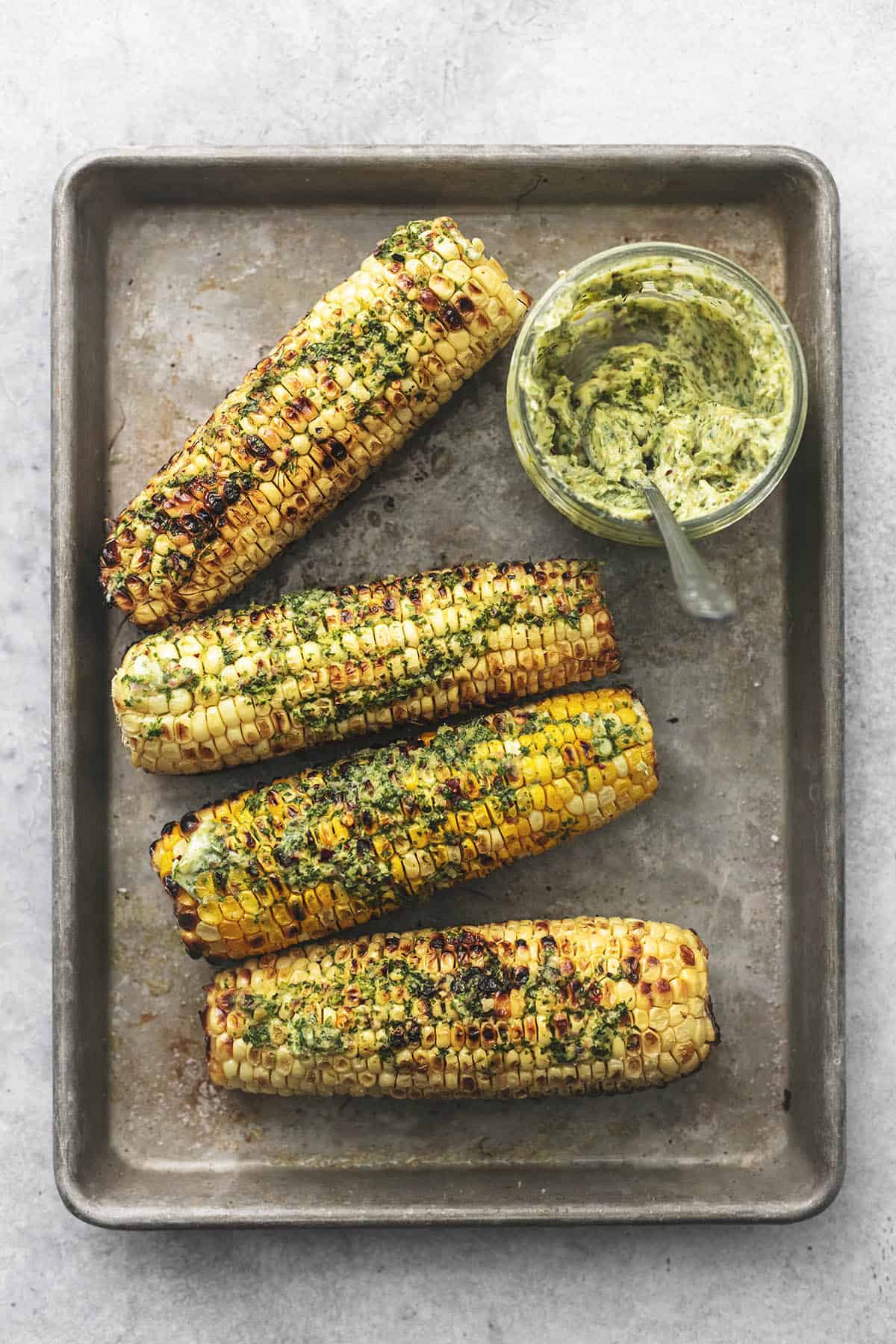 top view of grilled corn on the cob with chimichurri butter with a jar of more butter on the side all on a baking sheet.