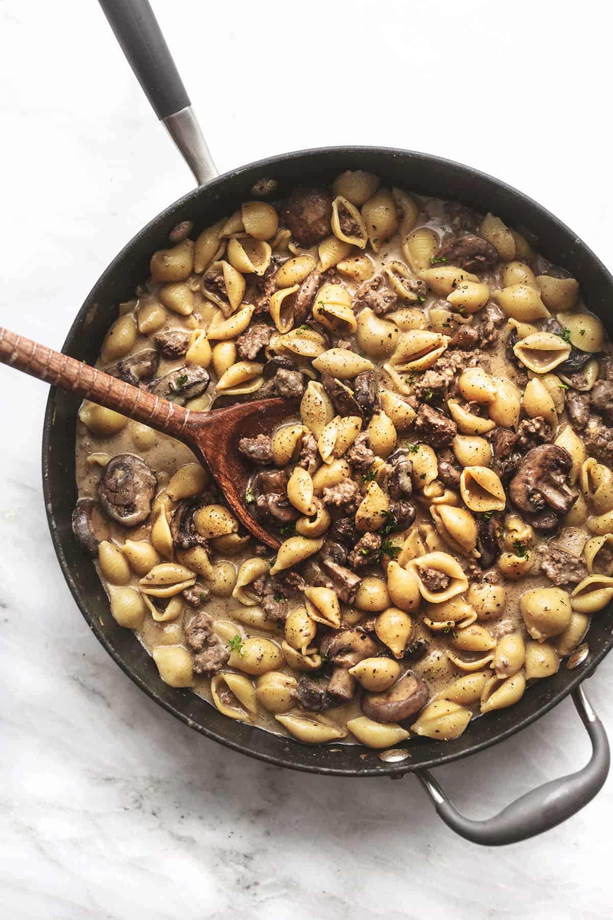 overhead view of ground beef stroganoff with pasta shells and serving spoon in black skillet on marble top