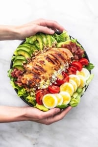hands holding bowl of salad with avocado, bacon, chicken, tomatoes, cheese, hard boiled eggs