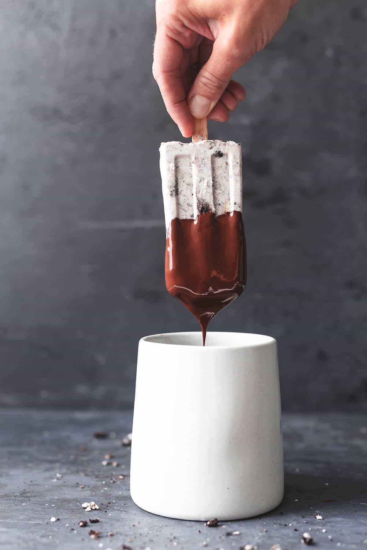 a hand dipping a cookies and cream pudding pop into a mug of melted chocolate.