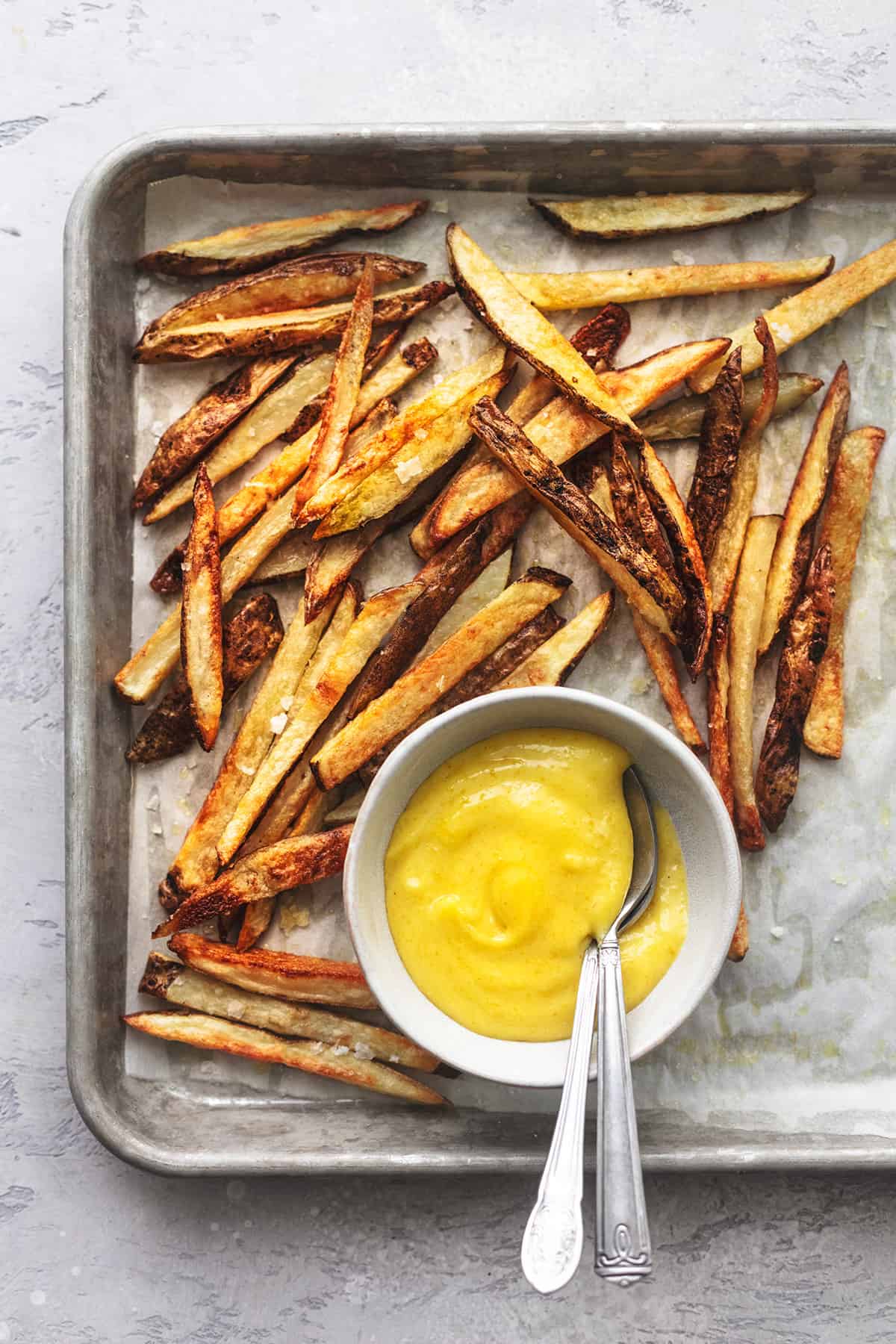 close up top view of baked french fries on a baking sheet with yellow sauce in a bowl.