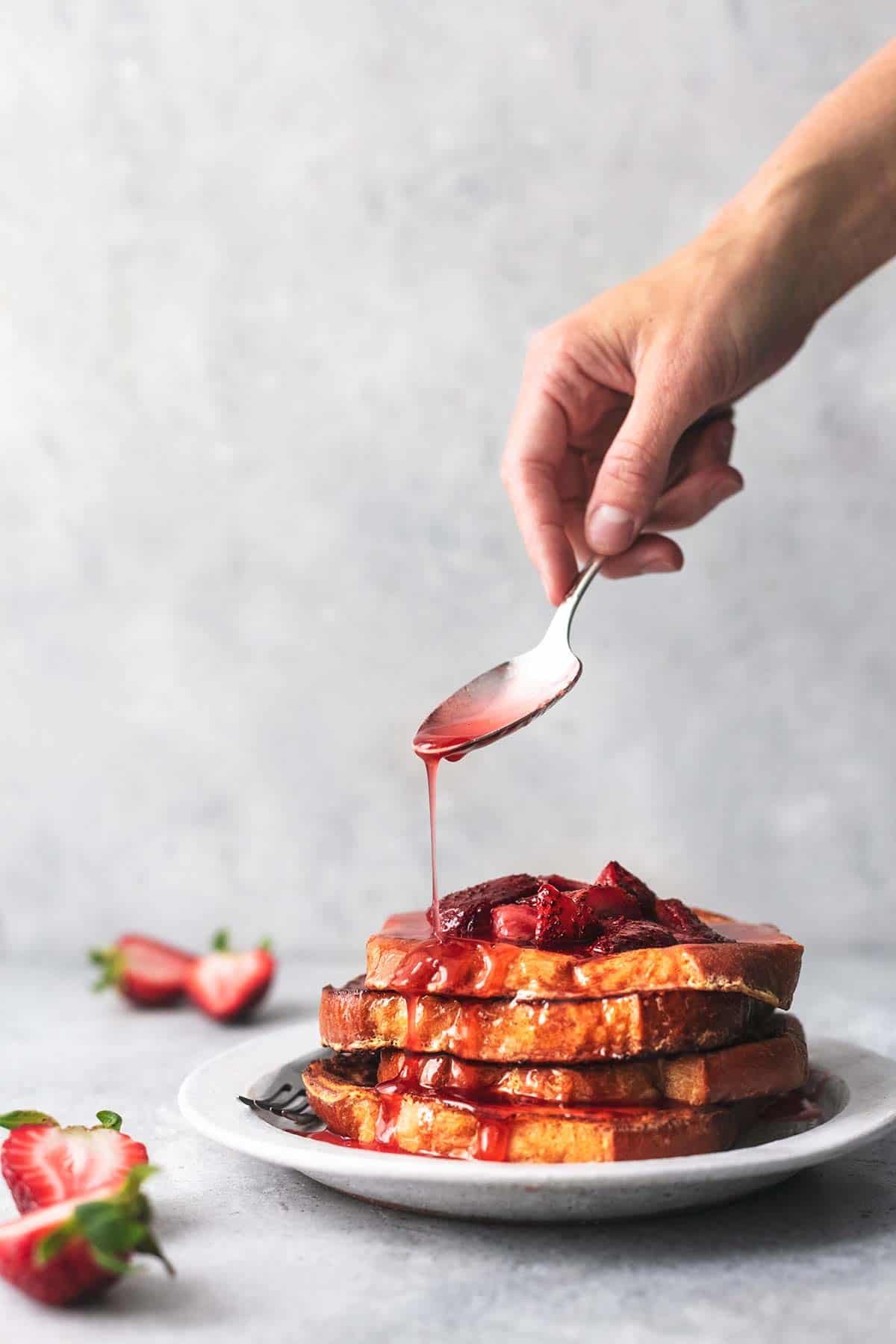 a hand drizzling strawberry sauce with a spoon onto a stack of strawberries and cream French toast on a plate.