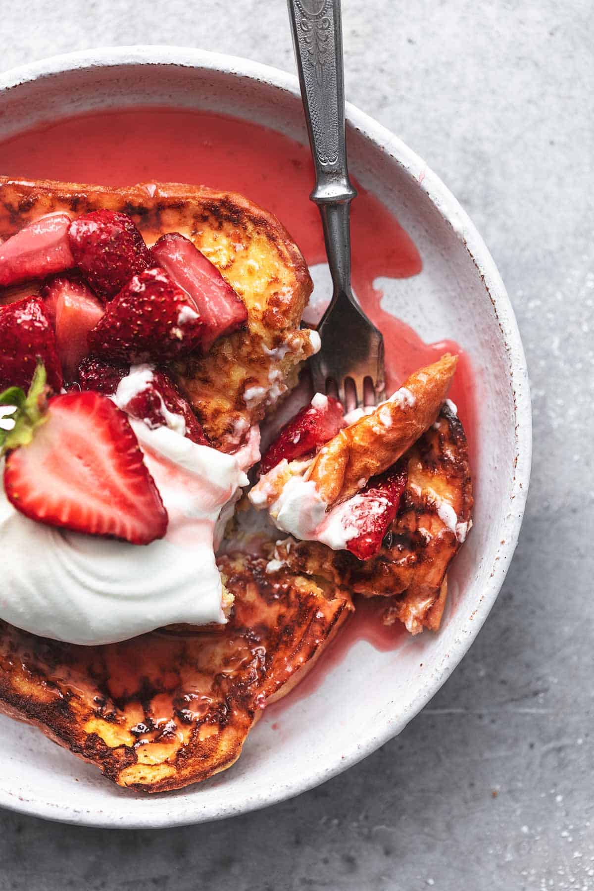 close up top view of strawberries and cream French toast topped with whipped cream, strawberries, and strawberry sauce with a bite on a fork.